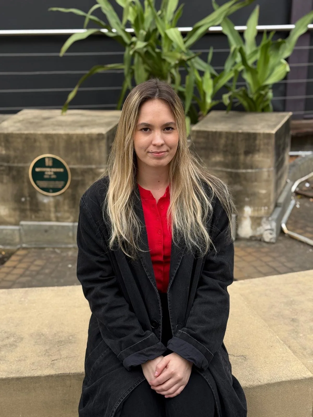 Young woman with long blonde hair sitting on a concrete bench outdoors, wearing a red shirt and black jacket, with large potted plants in the background.