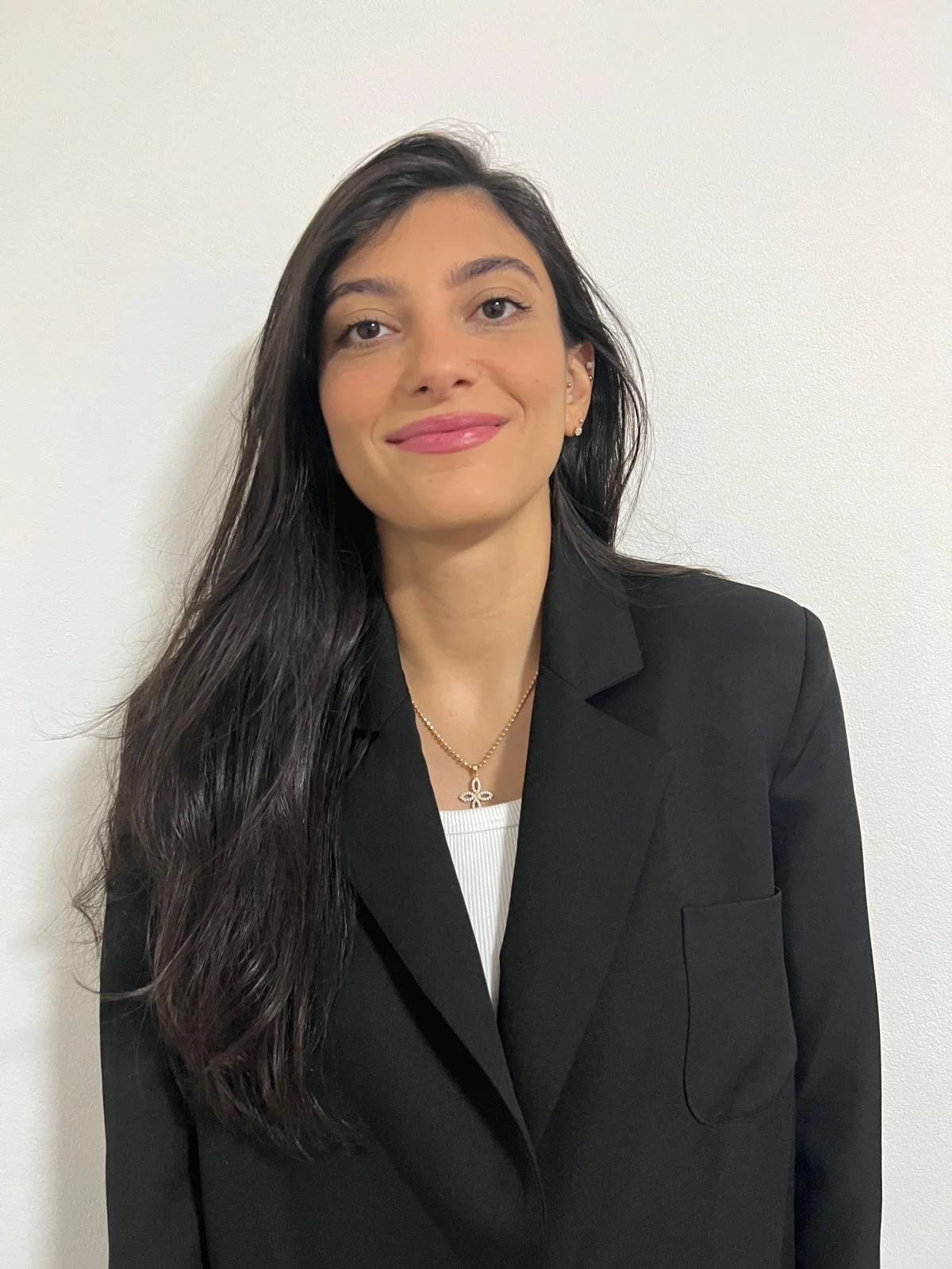 A woman with long dark hair, wearing a black blazer, white top, and gold jewelry, standing against a plain white wall.