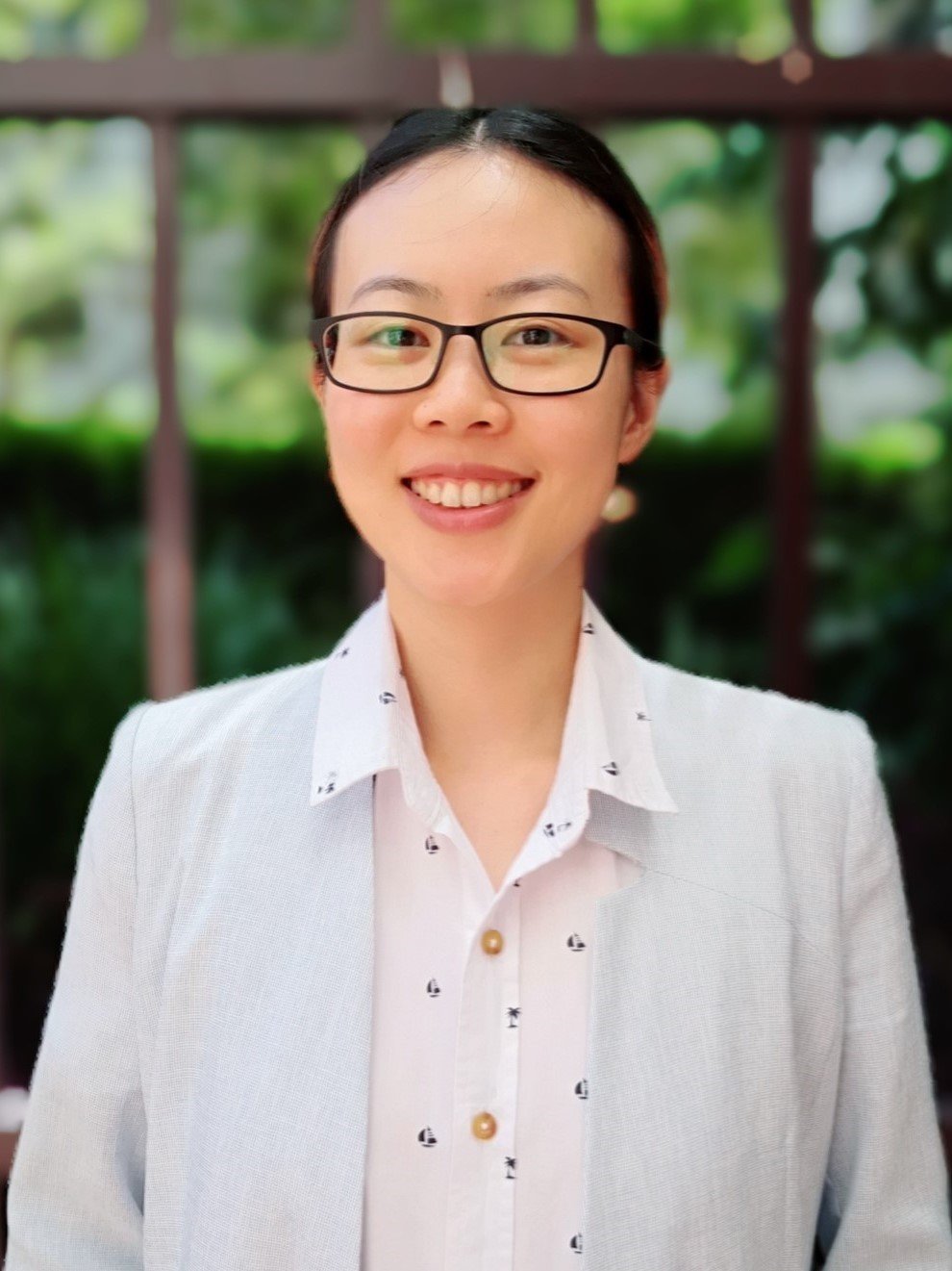 A young woman with glasses smiling outdoors, wearing a white blazer and a white shirt with small black sailboat patterns, with blurred greenery in the background.