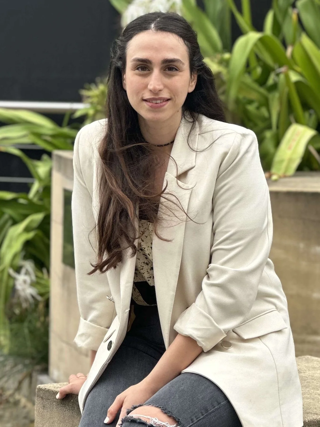 A woman with long dark hair seated outdoors near greenery, wearing a beige blazer over a patterned top and ripped black jeans.