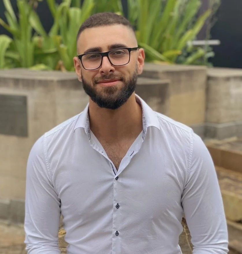A man with glasses, a beard, and short hair wearing a white button-up shirt, standing outdoors in front of green plants and concrete planters.