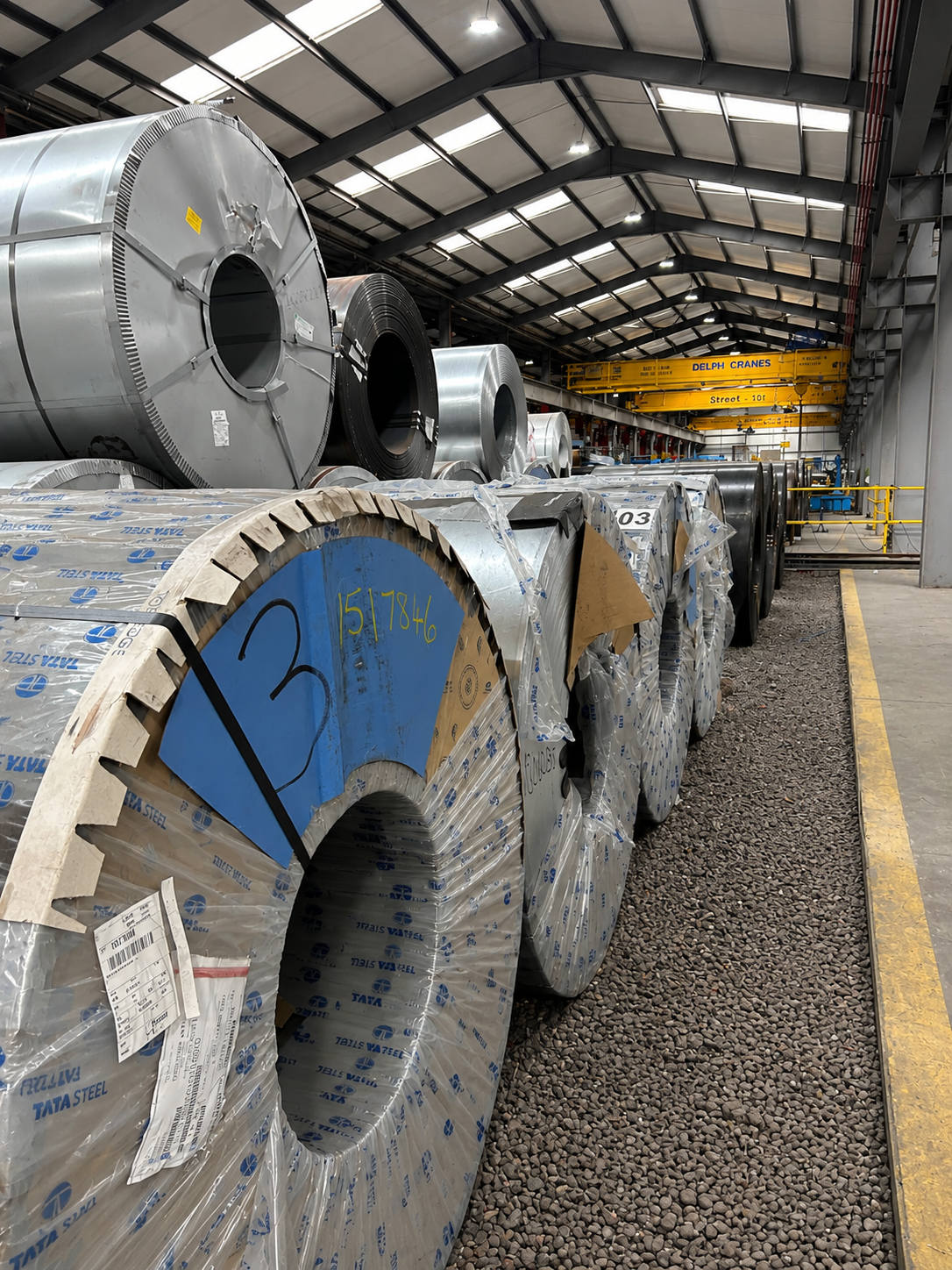 Metal coils stored inside a warehouse with a high ceiling, yellow overhead cranes, and a designated walking path on gravel and concrete floor.