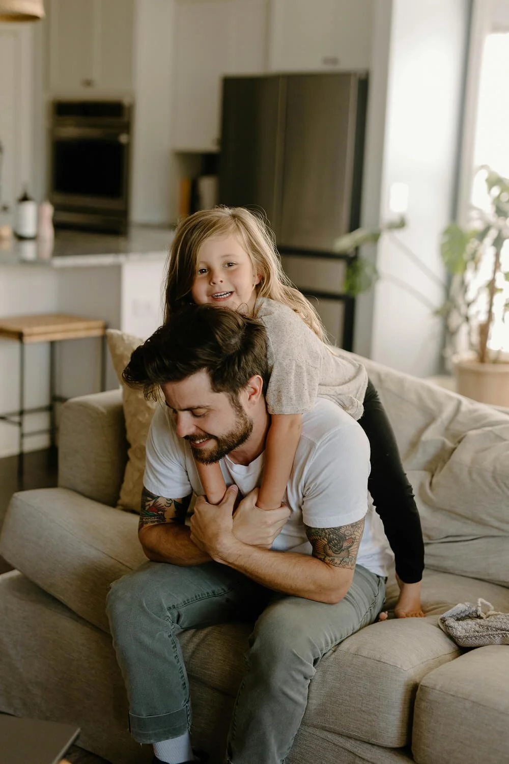 A man with tattoos sitting on a beige couch with a young girl on his shoulders, smiling and playing together in a bright, modern living room.