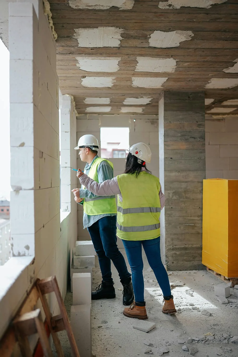 Two construction workers wearing helmets and safety vests on a building site, looking out the window and discussing construction plans.