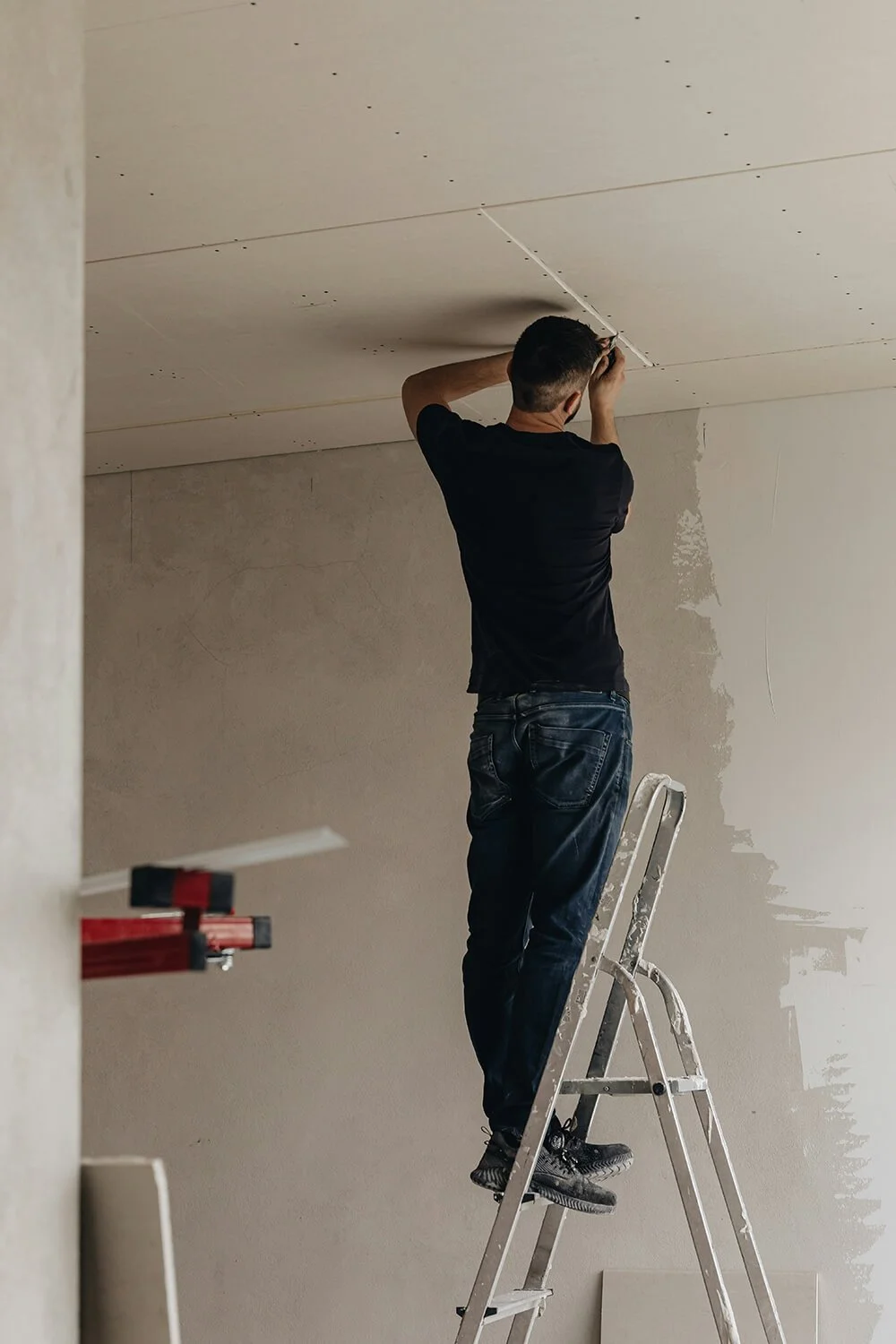A person standing on a ladder installing drywall on a ceiling in an unfinished room.