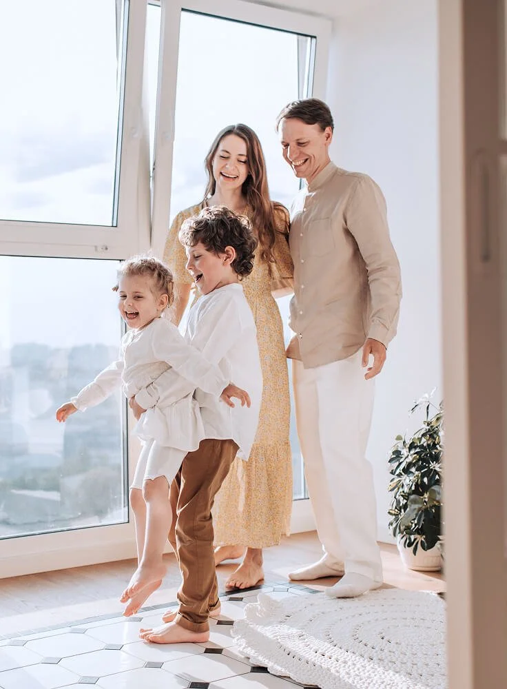 A family of four, including two children, a man and a woman, in a bright room with large windows, all smiling and playing together.