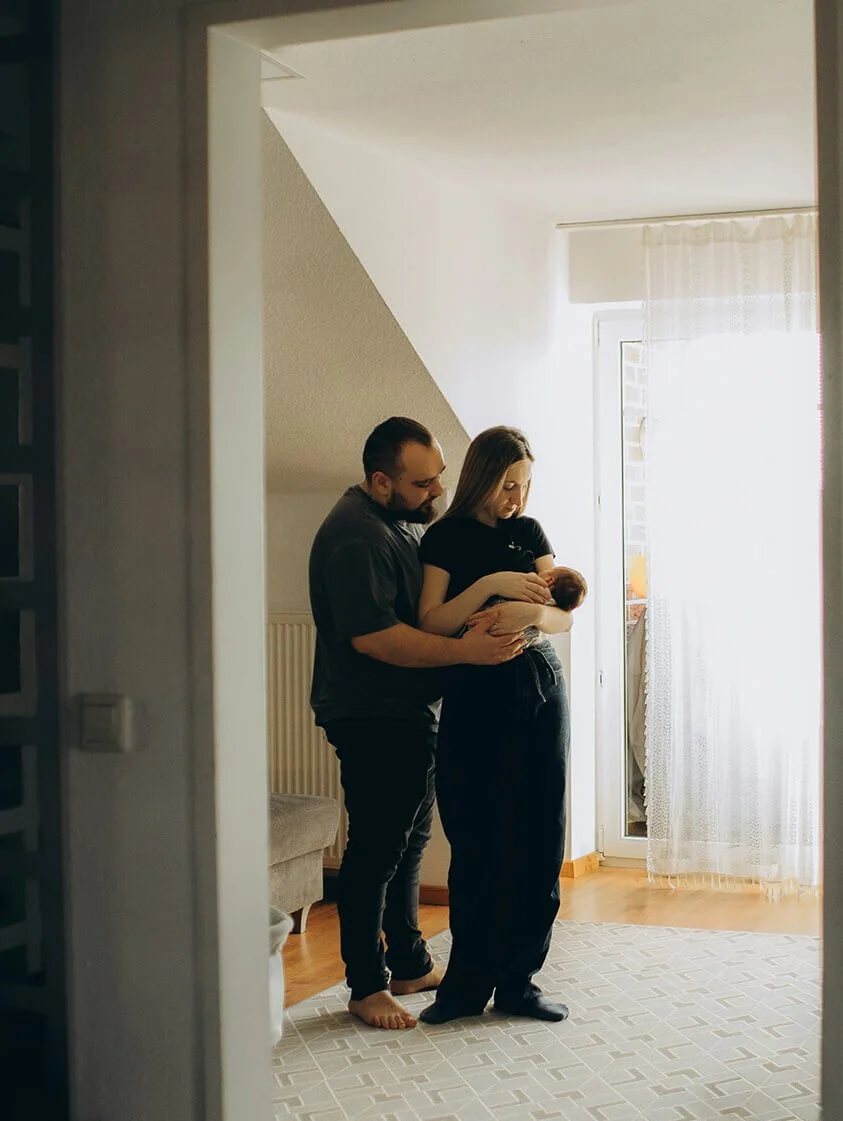 A couple with light skin, a man and a woman, are standing in a bright room with a door and a window with curtains. The woman is holding a newborn baby, and the man is gently touching the baby as they look at it.