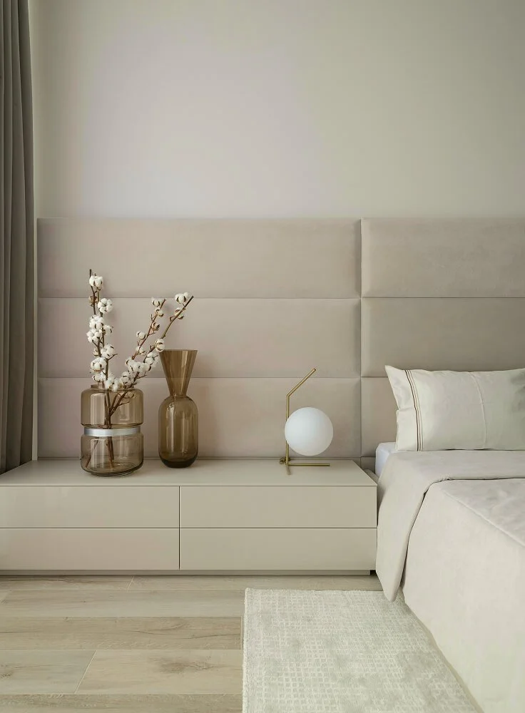 A bedroom with a beige upholstered headboard, beige curtains, and a beige dresser with three drawers. On the dresser are two amber-colored vases, one with cotton branches, and a modern table lamp with a white spherical shade and gold stand. Part of a bed with beige bedding and pillows is visible.