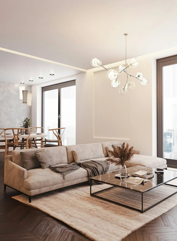 Modern living room with a beige sofa, a marble coffee table with a decorative plant, and large windows allowing natural light.
