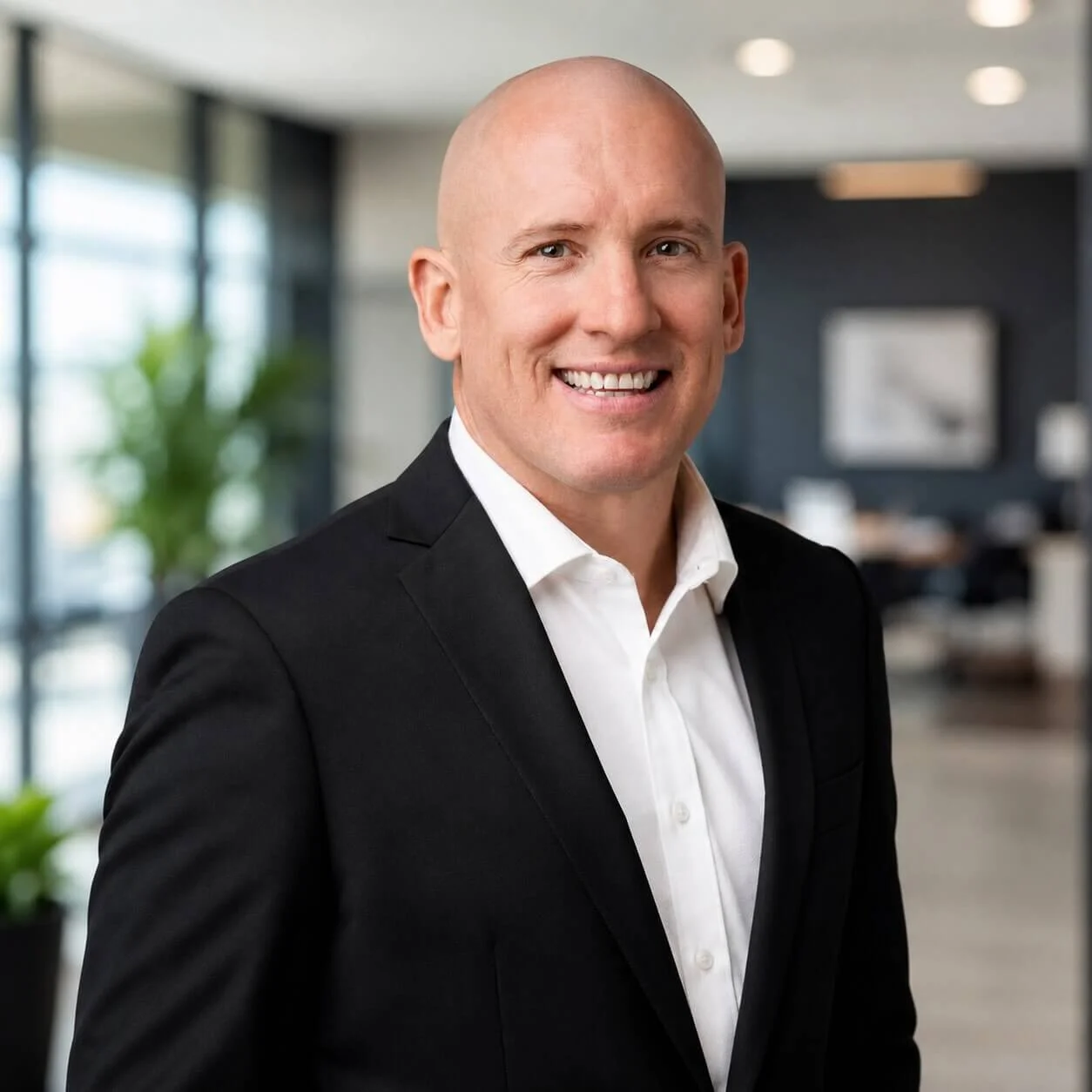 Portrait of a smiling, bald man in a black suit and white shirt, standing in a modern office with large windows and indoor plants in the background.