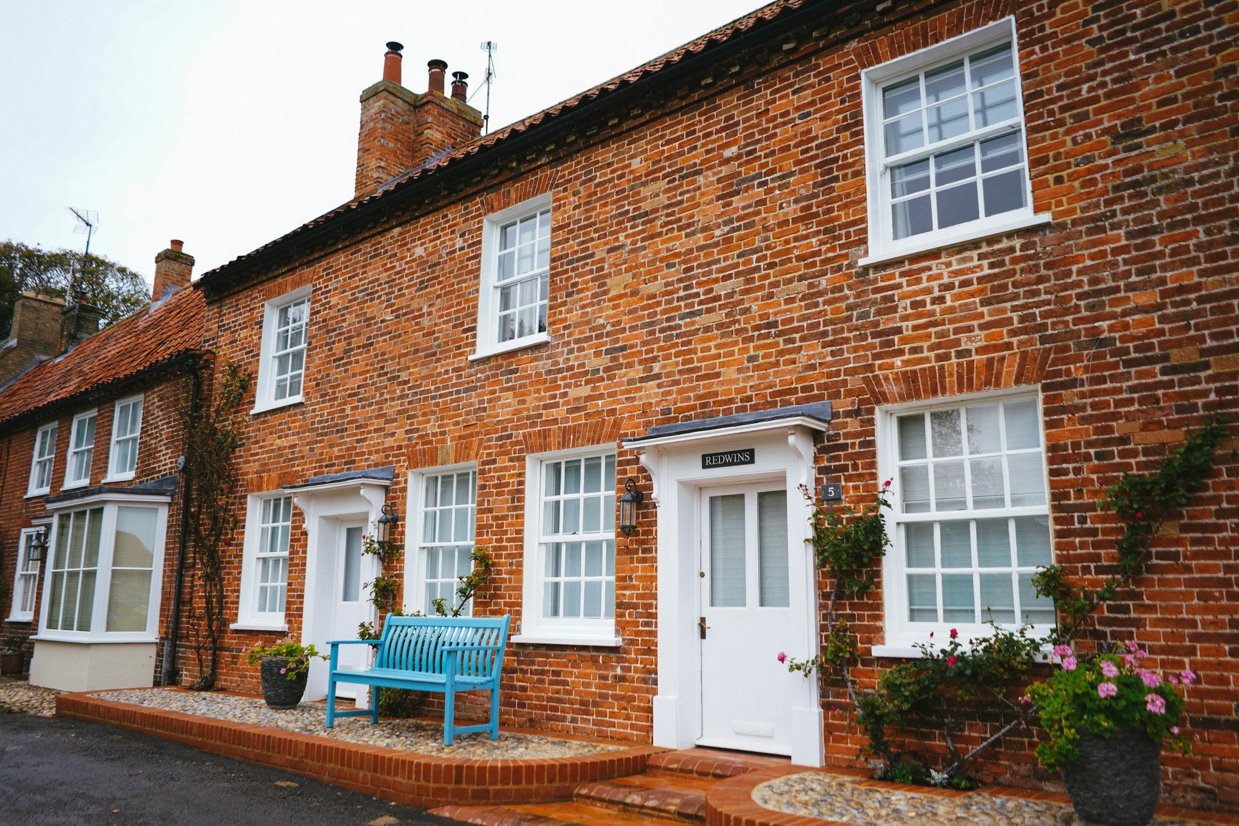Red brick townhouse with white framed windows, a white door with a black nameplate labeled "Redwings," blue bench, potted plants, and pink flowers outside.