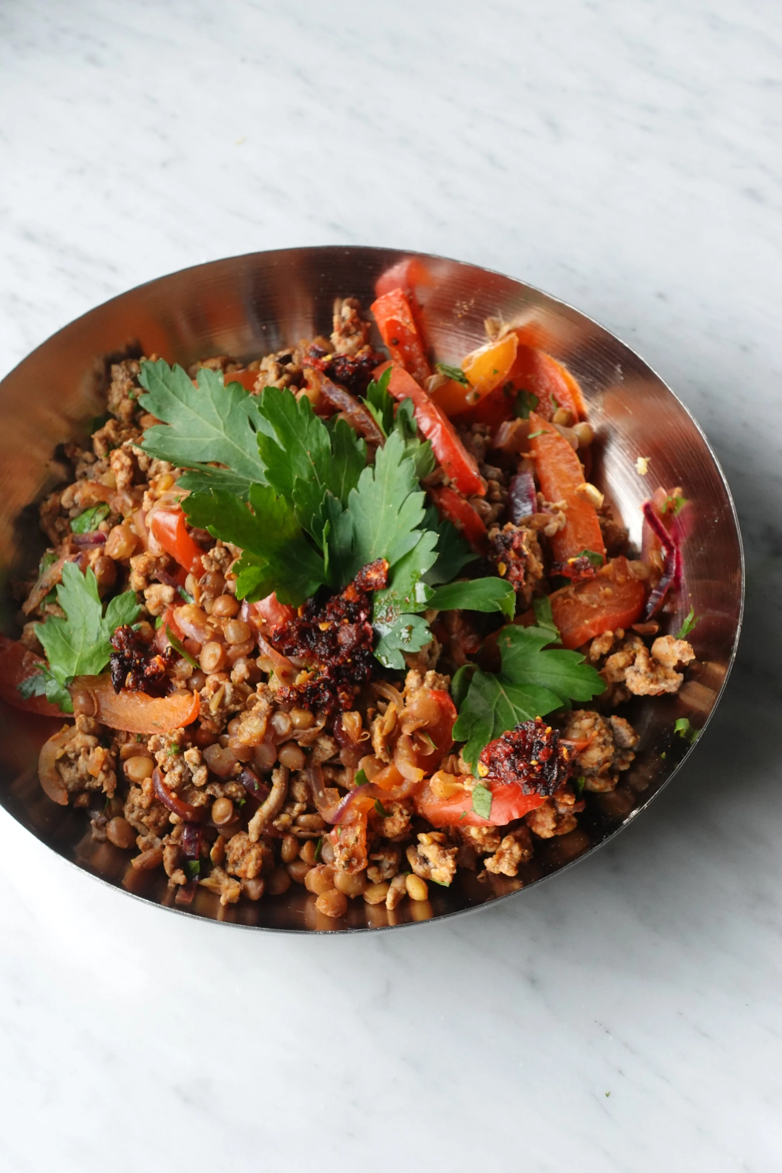 Een metalen kom met een salade van gehakt, linzen, gesneden tomaten, paprika, en verse peterselie, opgediend op een witte marmeren achtergrond.