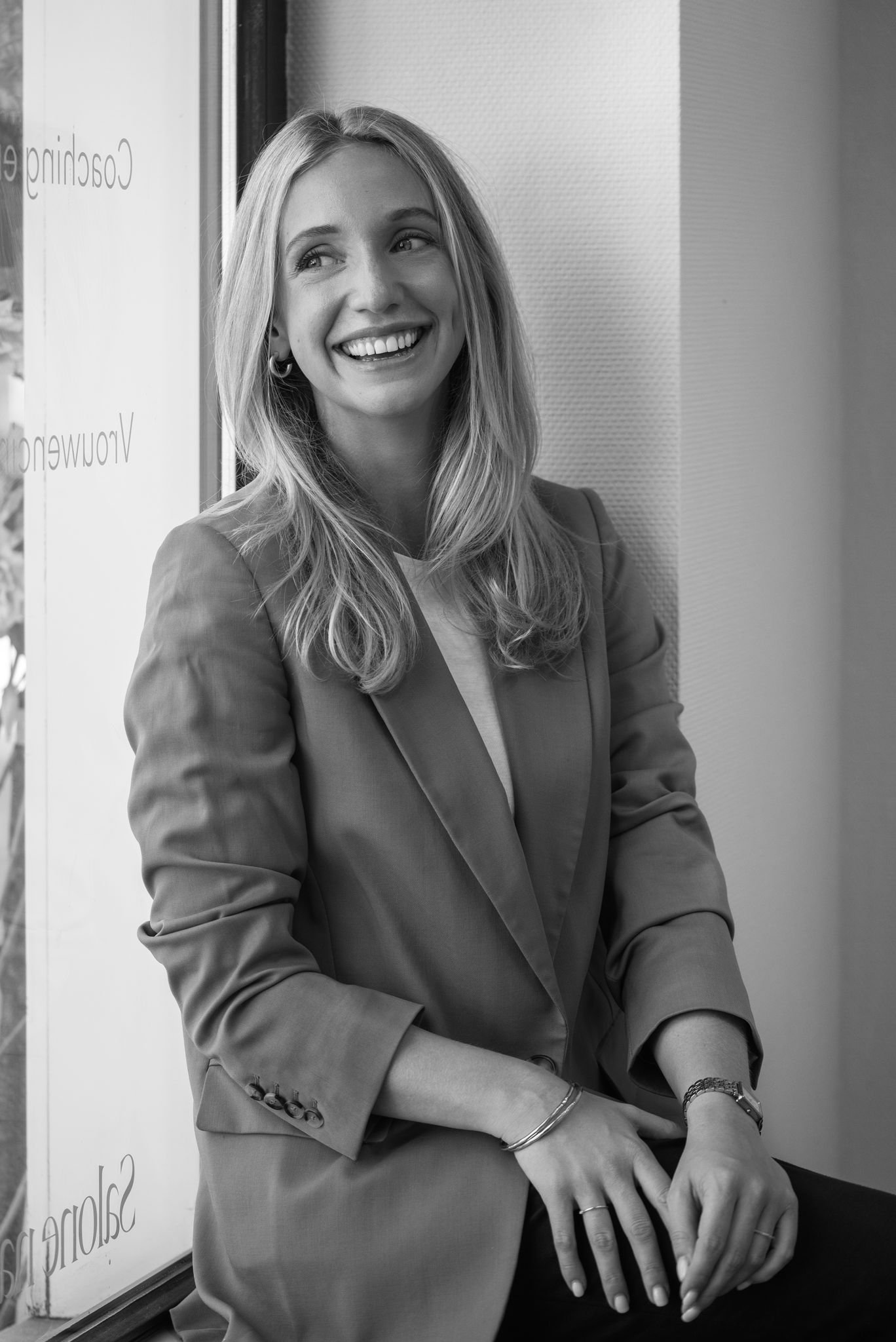 Zwarte en witte foto van een lachende vrouw met lang haar en een blazer, leunend tegen een muur, met een venster op de achtergrond.