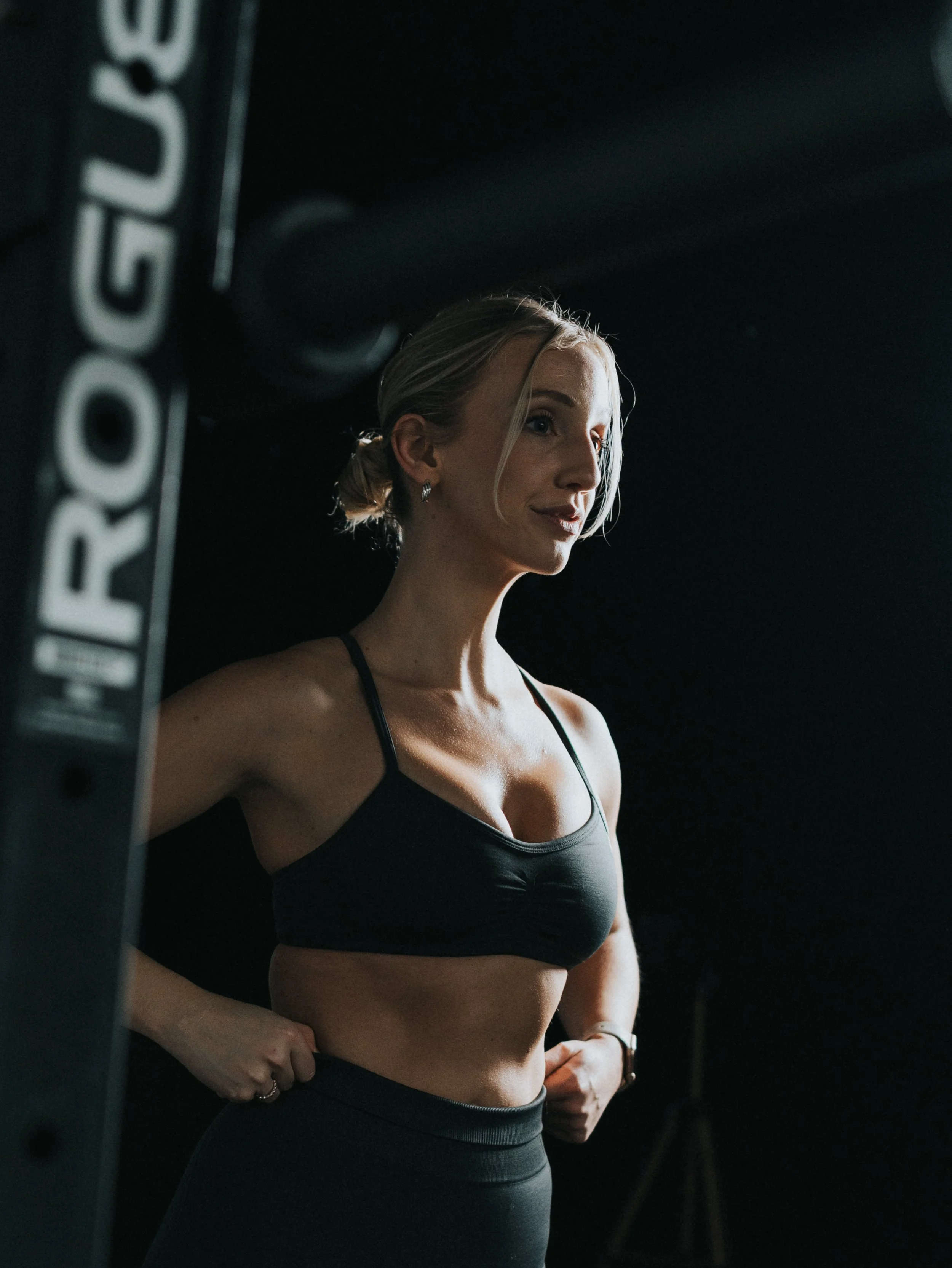Een vrouw met blonde haar in een paardenstaart draagt een zwart sports-bh en fitnessbroek, staat in een donkere omgeving en lijkt klaar voor een workout.