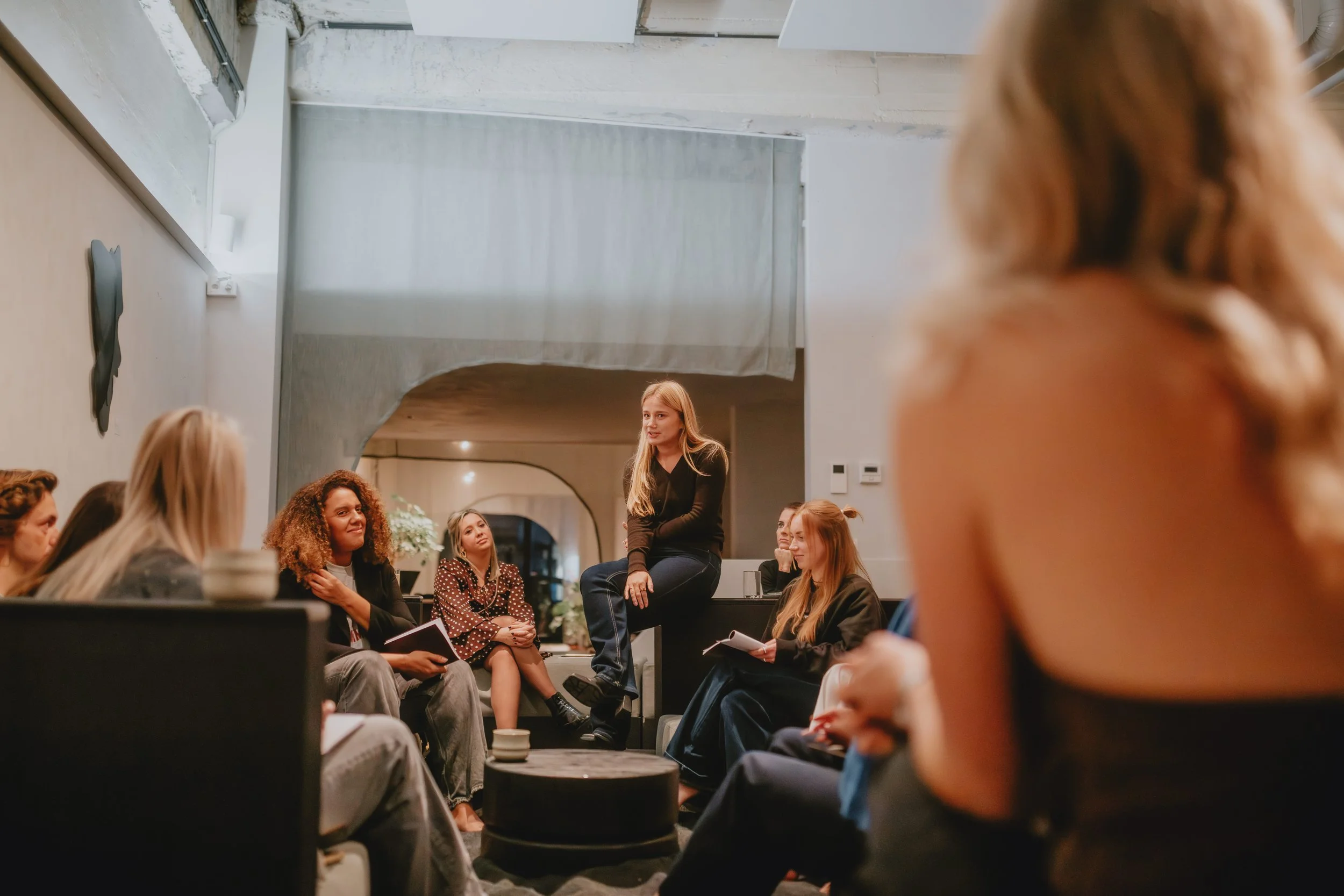 Een groep mensen zit in een modern interieur, terwijl een vrouw in zwart zit op een verhoging en spreekt. De aanwezigen luisteren aandachtig en nemen notities.