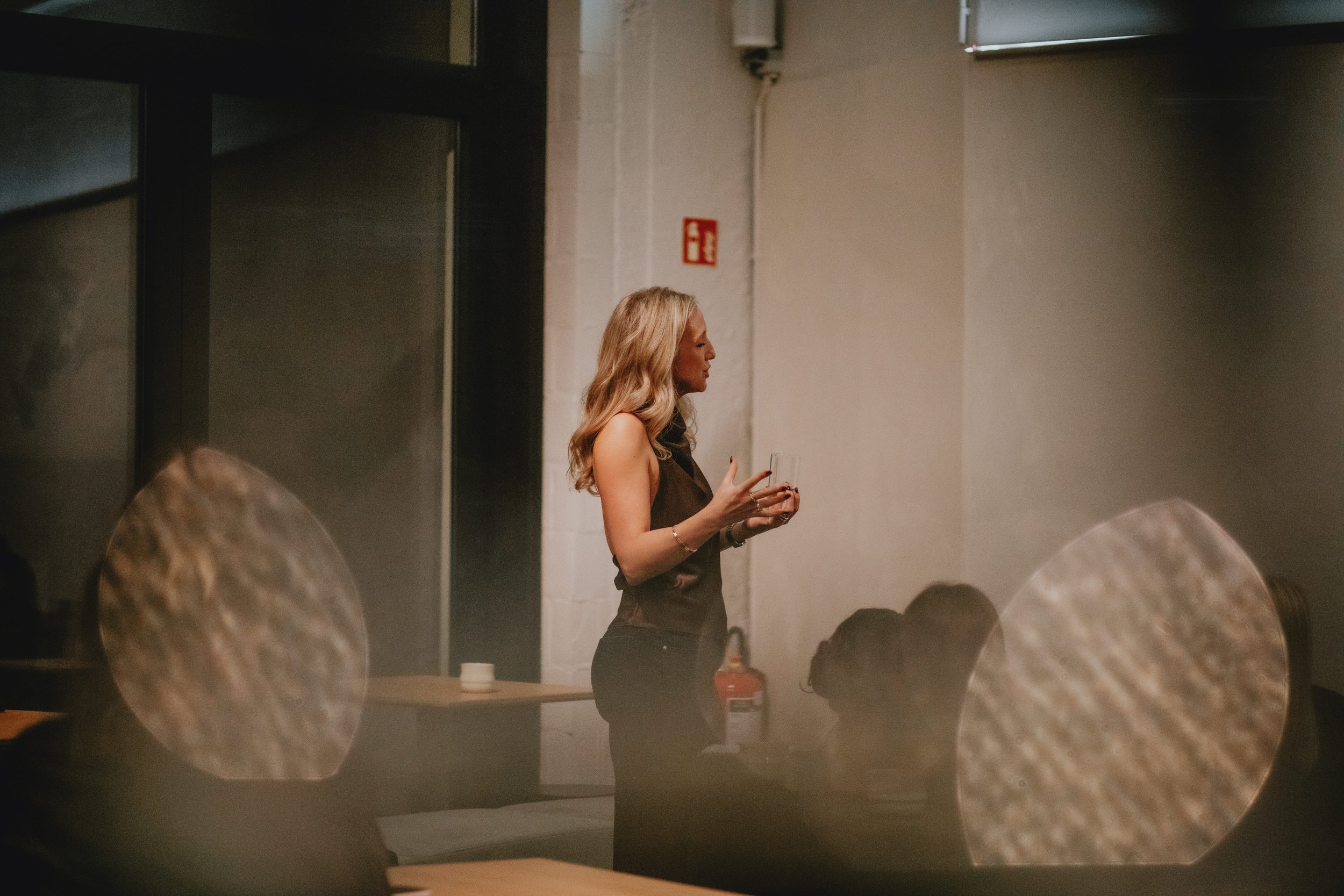 Vrouw met lange blonde haar die staat en een glas vasthoudt, binnen in een ruimte met witte muren, met enkele mensen die op de voorgrond zitten.