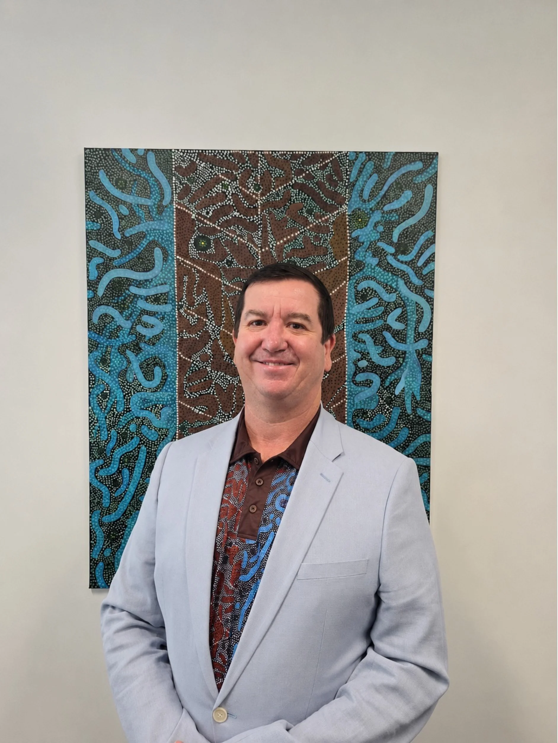 A man with short dark hair smiling, wearing a light grey suit and a patterned shirt, standing in front of a colourful abstract painting featuring blue, brown, and black designs with white dots on a white wall.