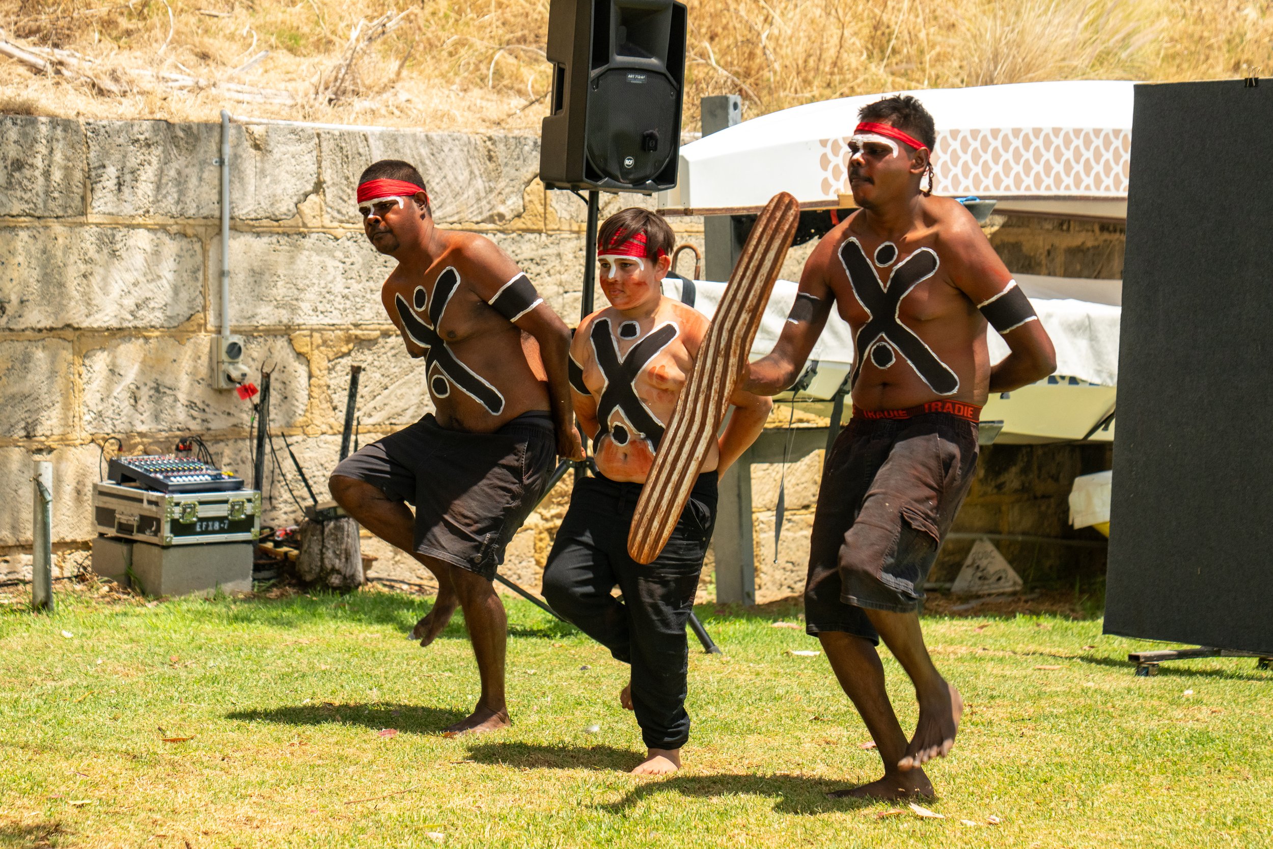 Three men with body paint and red headbands perform a traditional dance outdoors on grass, with a stone wall and musical equipment in the background.