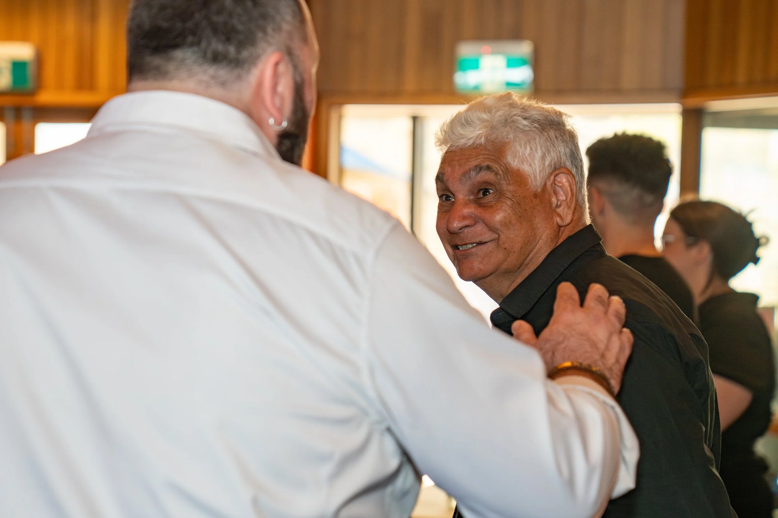 Two men dancing together, holding hands, during a social event in a room with wood-paneled walls and windows.