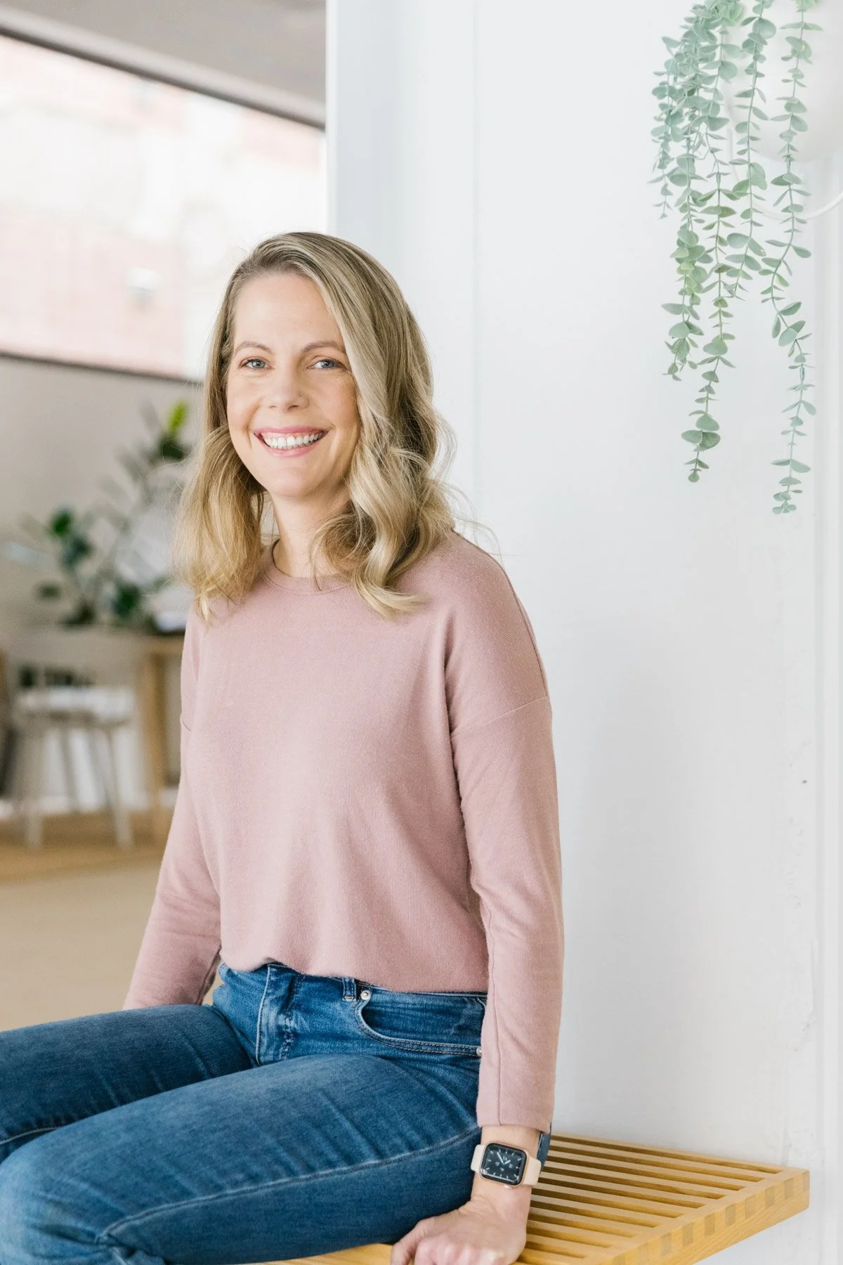 A woman with blonde hair sitting cross-legged on a wooden bench indoors, smiling at the camera. She's wearing a pink long-sleeve top, blue jeans, and an Apple Watch. The background includes a window, some potted plants, and a white wall with hanging greenery.