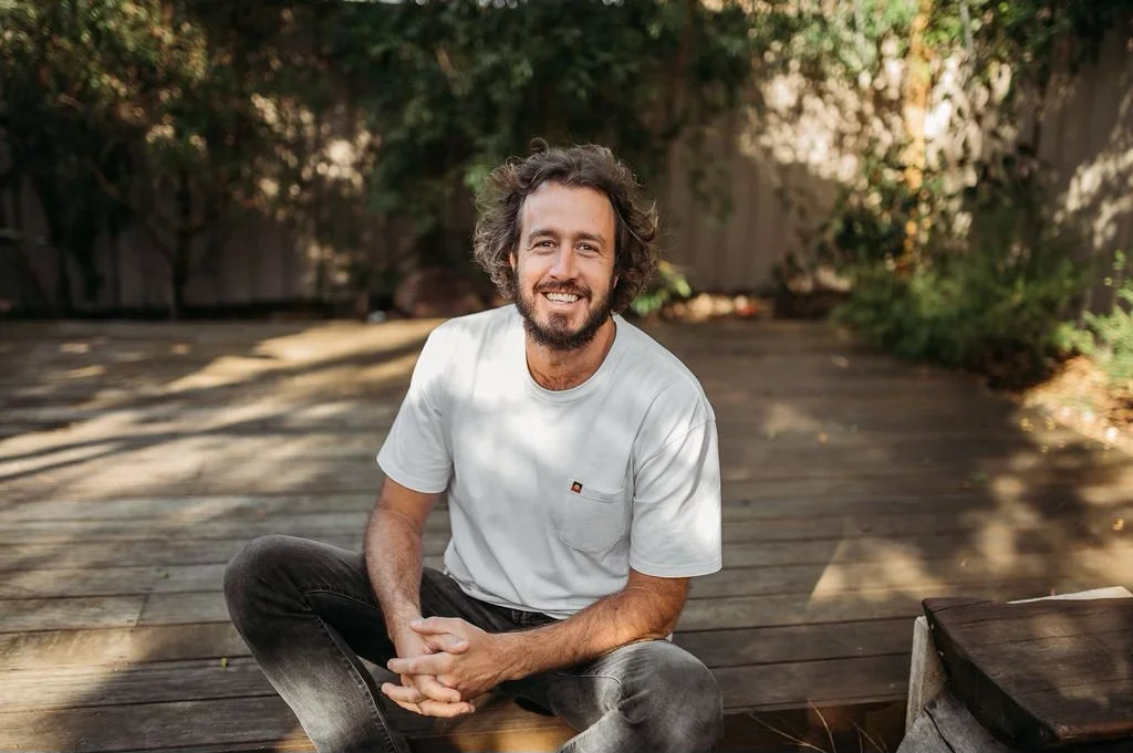A man with curly hair and a beard sitting cross-legged on a wooden deck, smiling at the camera, with trees and a fence in the background.