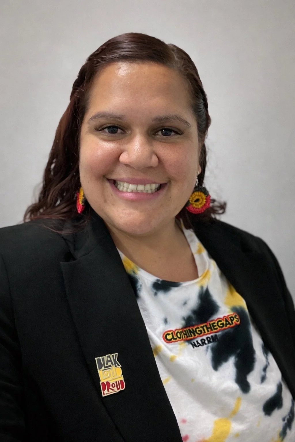 Smiling woman with brown hair tied back, wearing a black blazer with pins, colourful earrings, and a tie-dye T-shirt that says 'Clothing the Gaps' and 'NARROW,' standing against a plain wall.