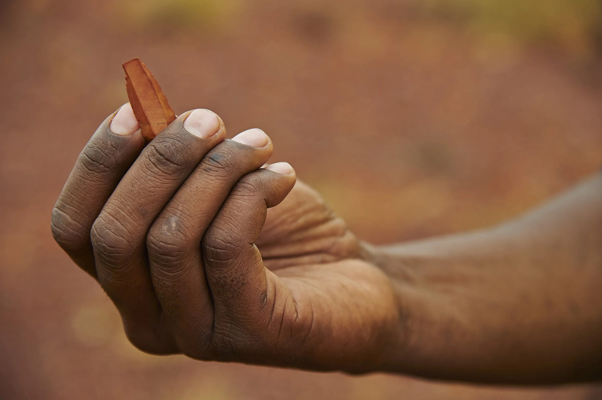 Close-up of a person's hand holding a small, rectangular, orange-brown object with fingers and thumb visible, against a blurred reddish-brown background.
