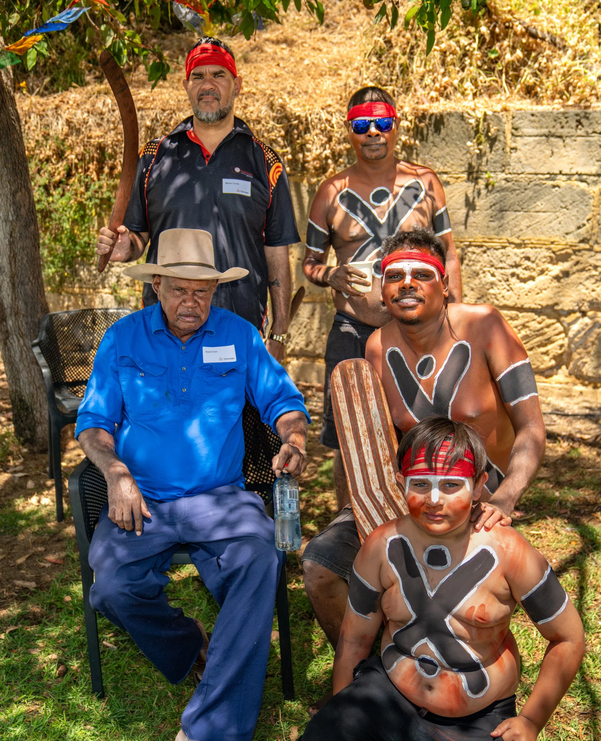 Group of five men, some with painted faces and body art, gathered outdoors, posed for a photo with some standing and some sitting; one man seated on a chair holding a water bottle and wearing a cowboy hat, others standing around him.