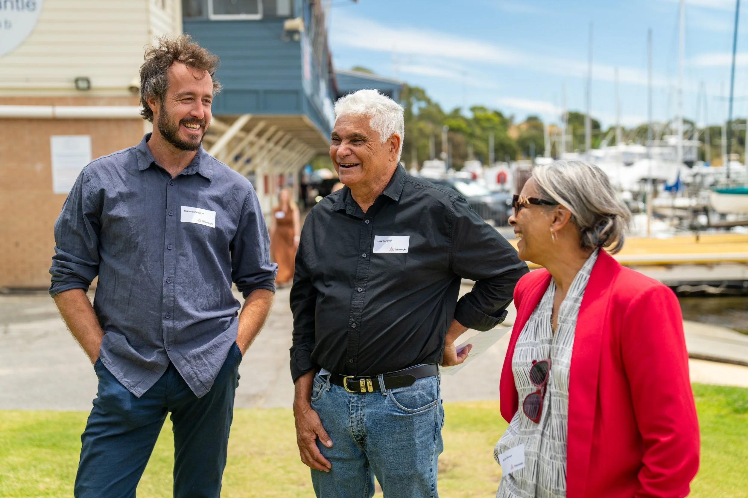 Three people, two men and one woman, are standing outdoors near a marina, engaging in conversation and smiling. The background includes boats and buildings.