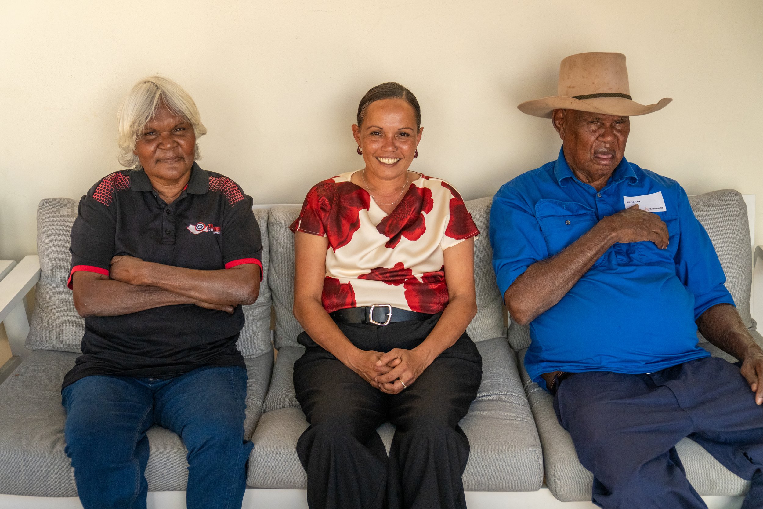 Three people sit on a gray sofa against a cream-colored wall, featuring two women and one man. The woman on the left has short, white hair and is wearing a black polo shirt with red details. The woman in the middle has her brown hair tied back and is wearing a blouse with a red floral pattern. The man on the right has a dark complexion, wears a large beige hat and a blue shirt, and has a name tag. The man appears to be looking at the camera, the woman in the middle is smiling, and the woman on the left has her arms crossed.