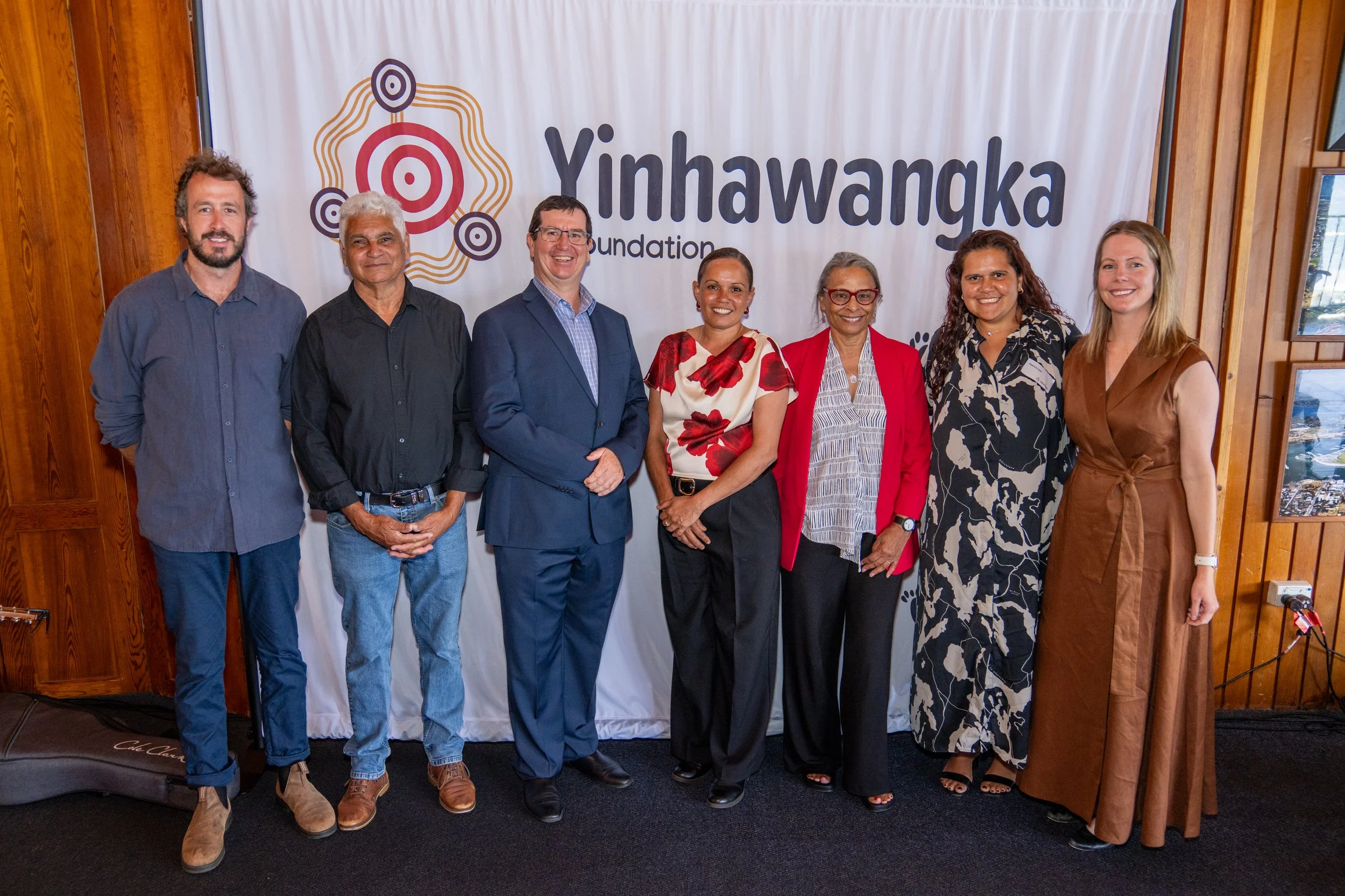 Group of seven diverse people standing in front of a Yinhawangka Foundation banner at an indoor event.