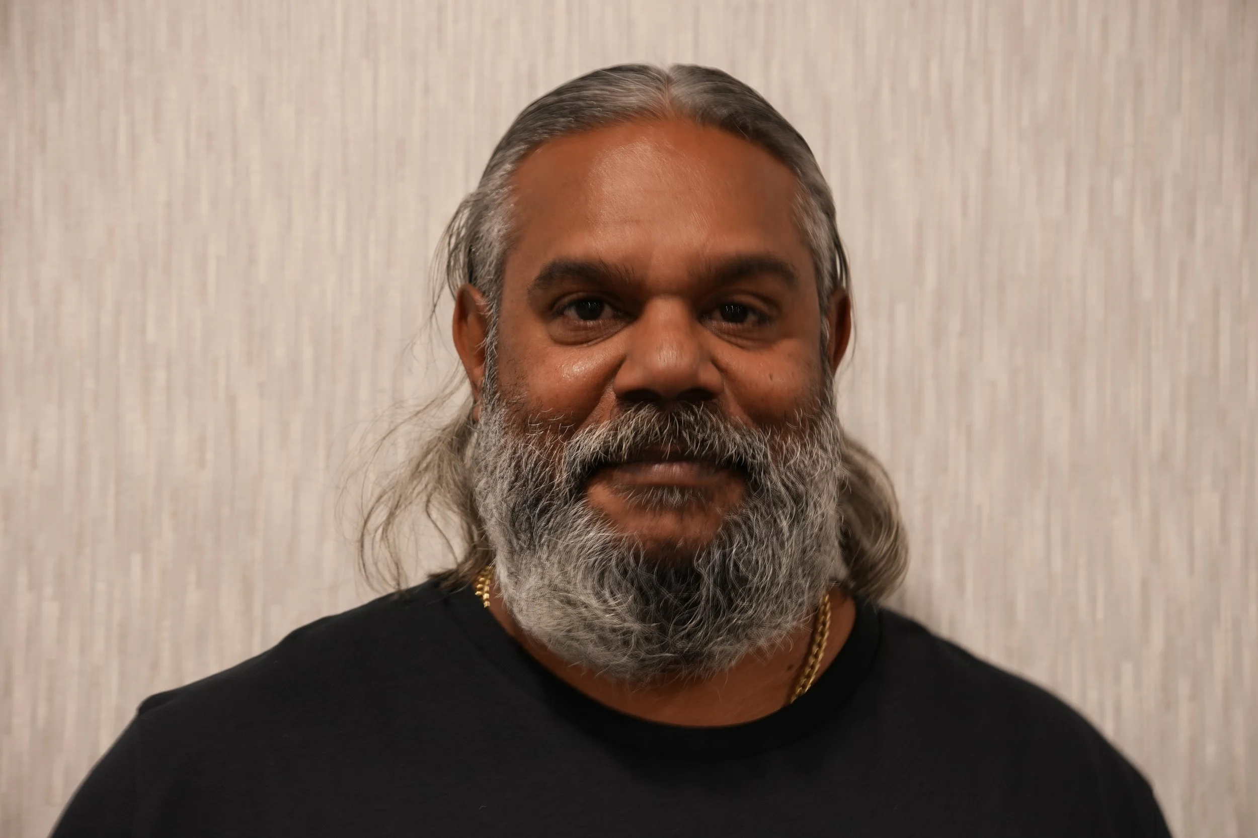 A close-up of a middle-aged man with grey hair and a beard, wearing a black shirt and a gold chain, standing in front of a light-coloured textured wall.