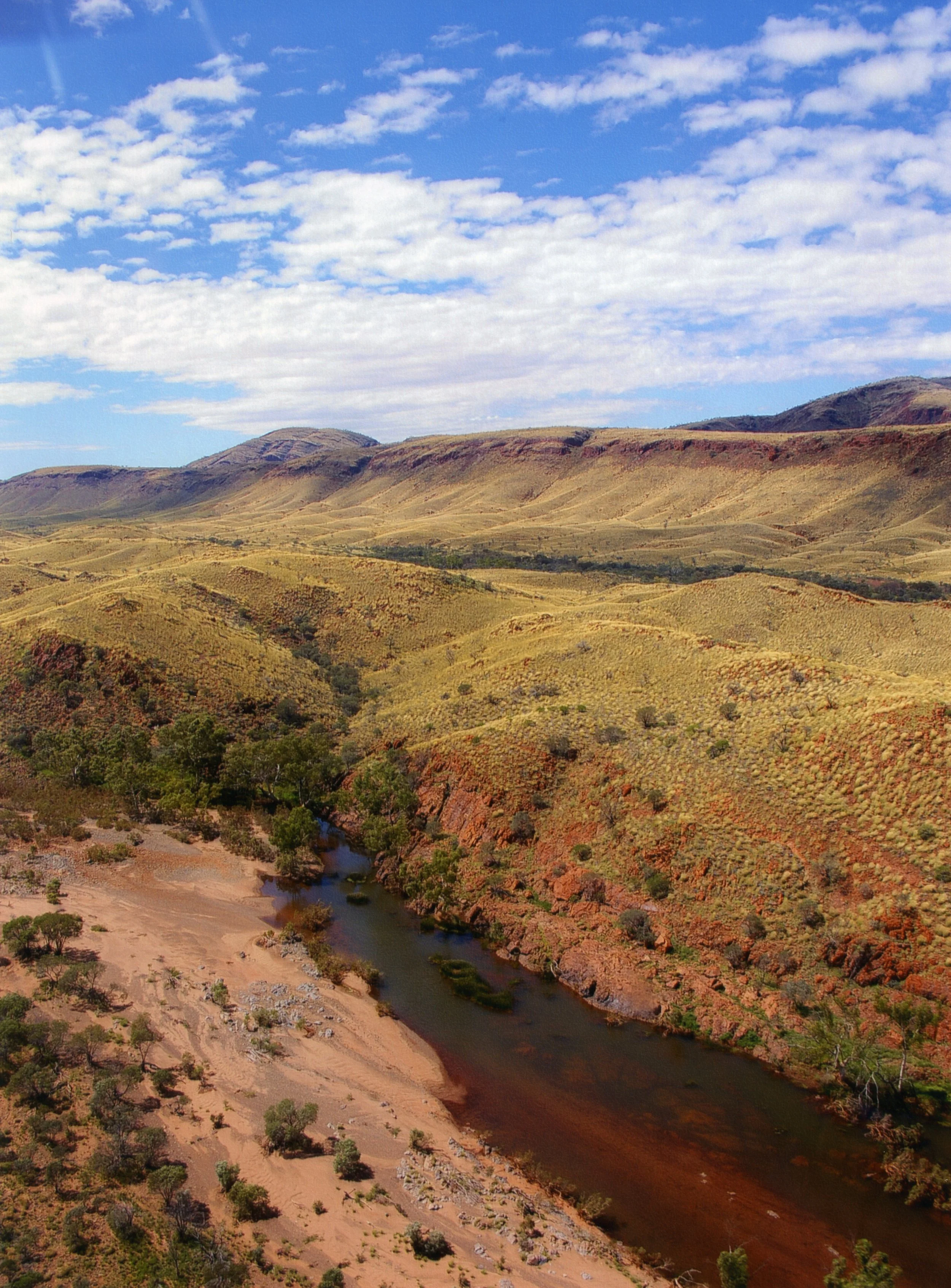 A landscape of rolling hills with sparse vegetation, a river flowing through the valley, and a partly cloudy sky overhead.