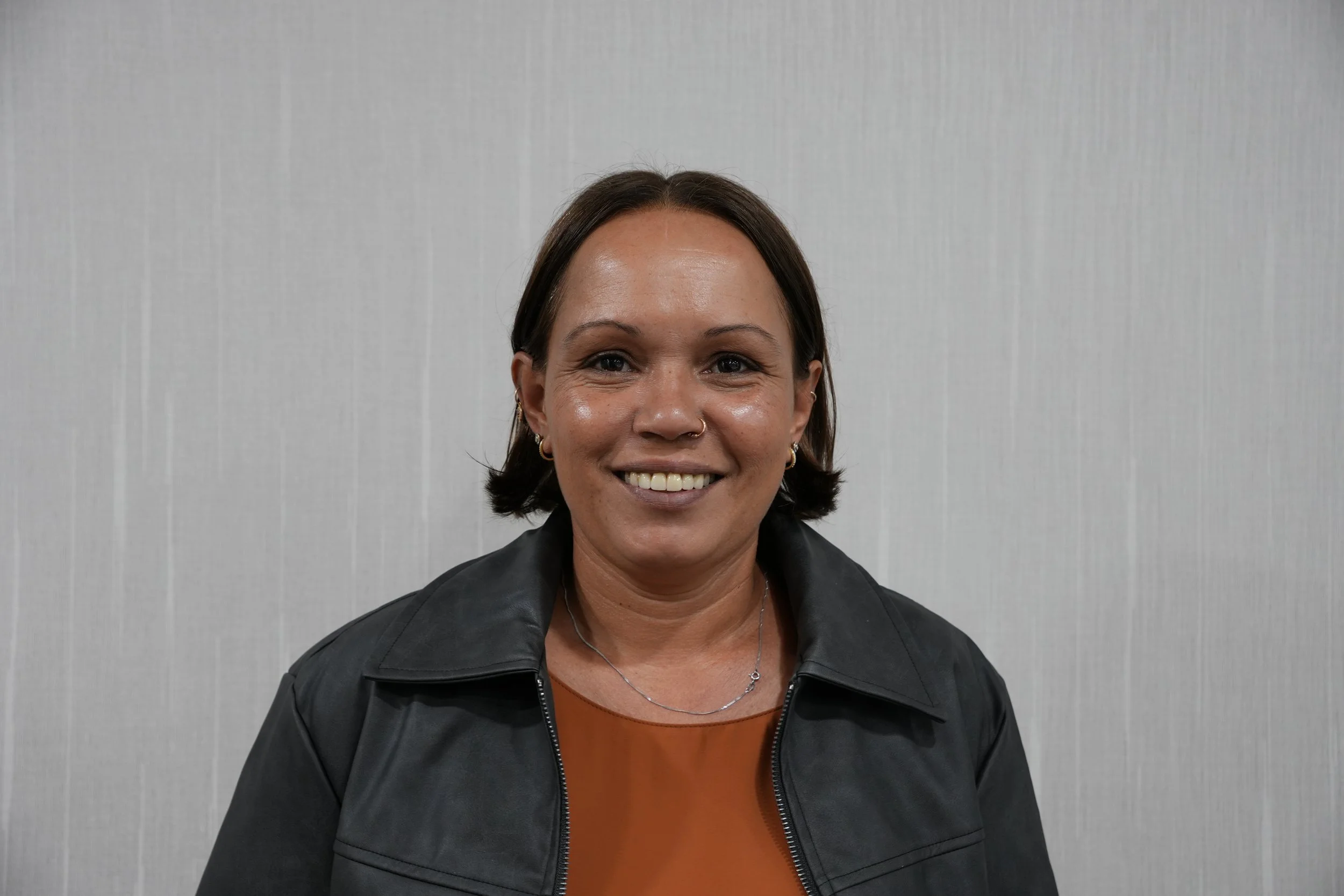 A woman with short dark hair, wearing earrings, a nose ring, an orange top, and a black leather jacket, smiling in front of a light-coloured wall.