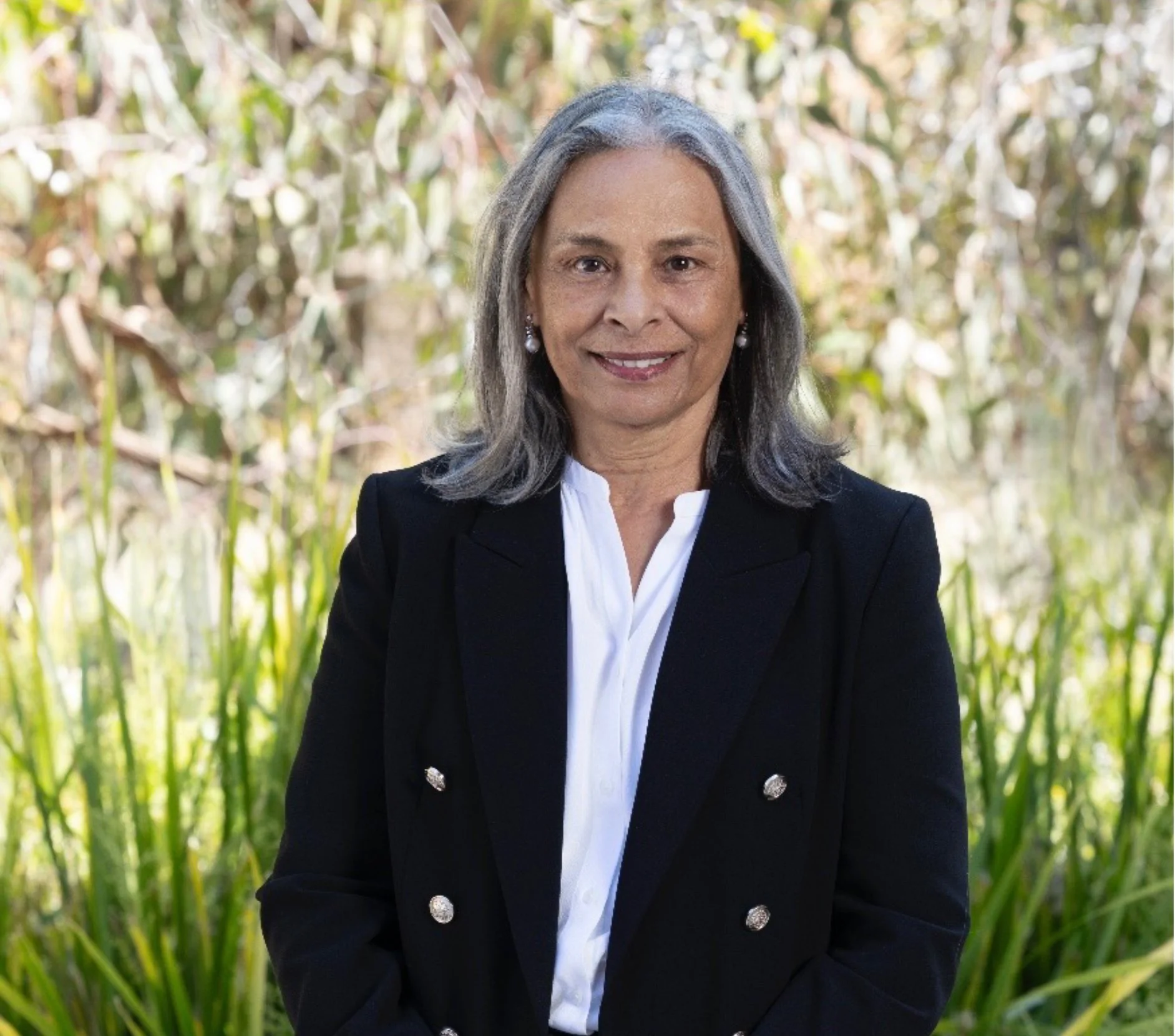 Portrait of a middle-aged woman with long gray hair, wearing a black blazer with silver buttons and a white shirt, standing outdoors with green foliage in the background.