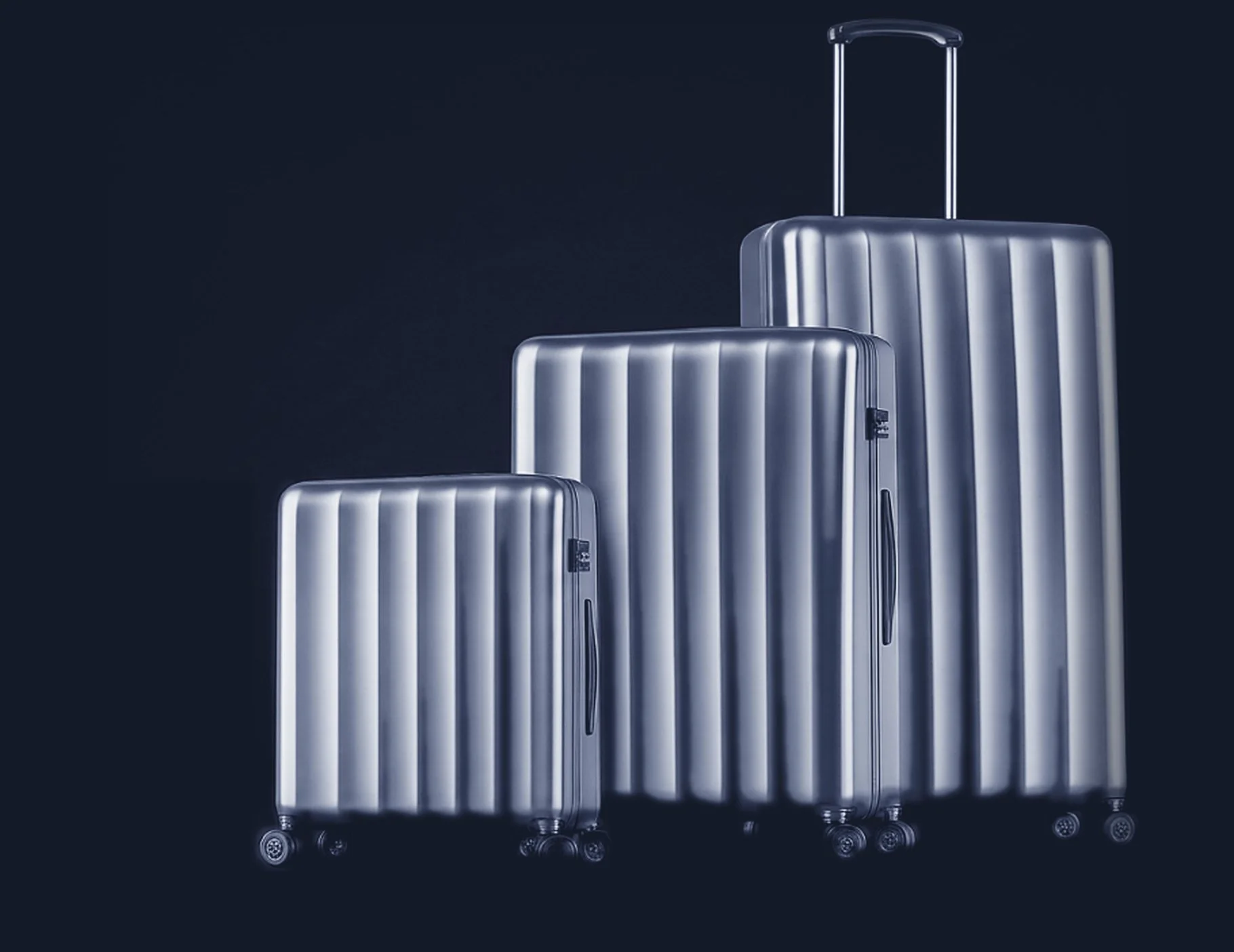 Three silver suitcases of varying sizes with horizontal ridges and retractable handles, set against a dark background.