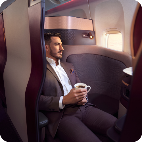Man in a business suit sitting in airplane seat, holding a coffee cup, looking out the window.