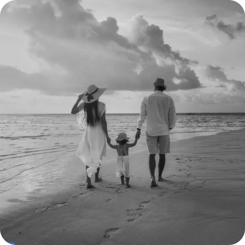 A family of three, consisting of a woman, a man, and a young girl, walking hand-in-hand on the beach during sunset, with clouds in the sky.