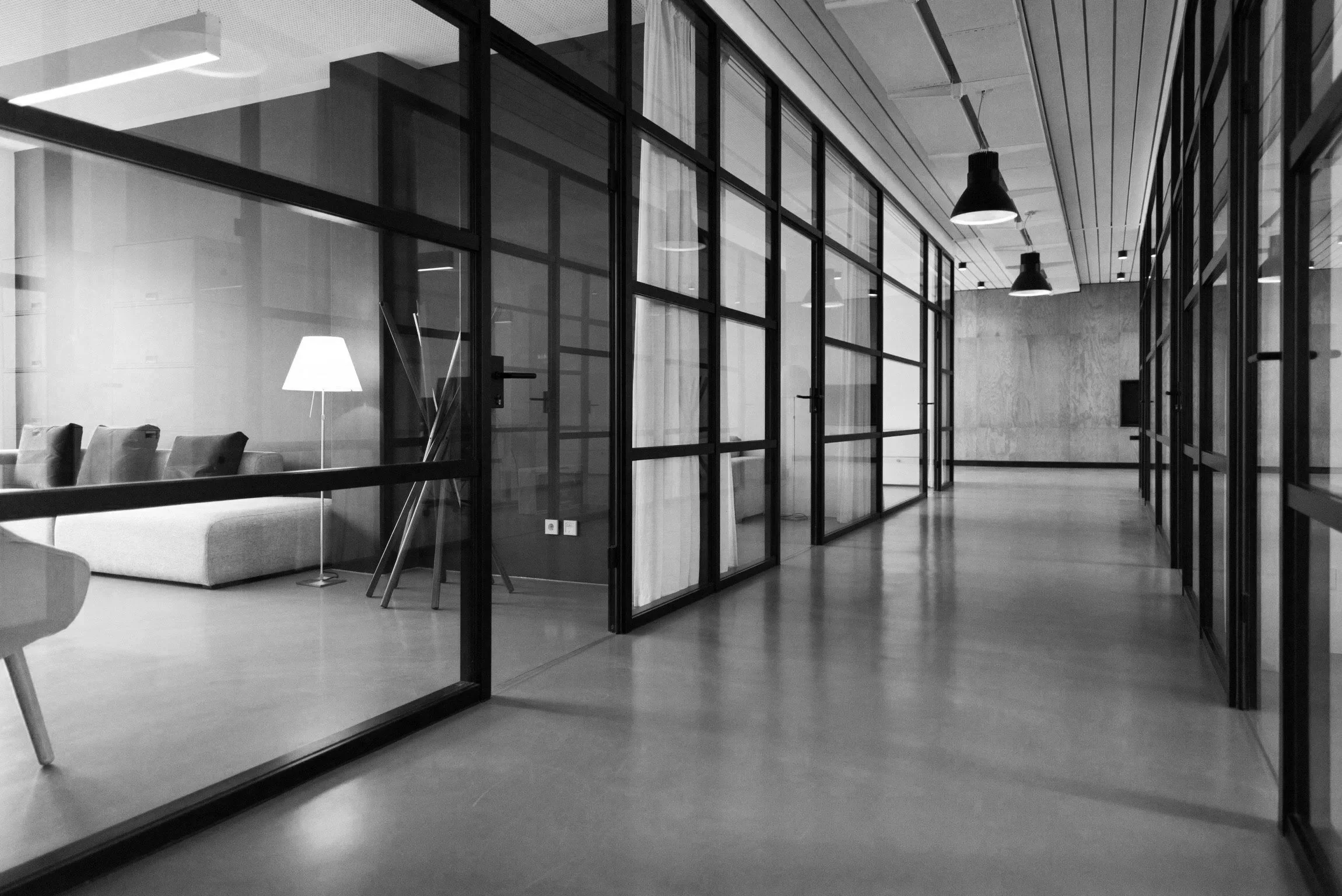 Black and white photo of an office space with glass walls, a seating area with chairs and a lamp, and a hallway with pendant lights.