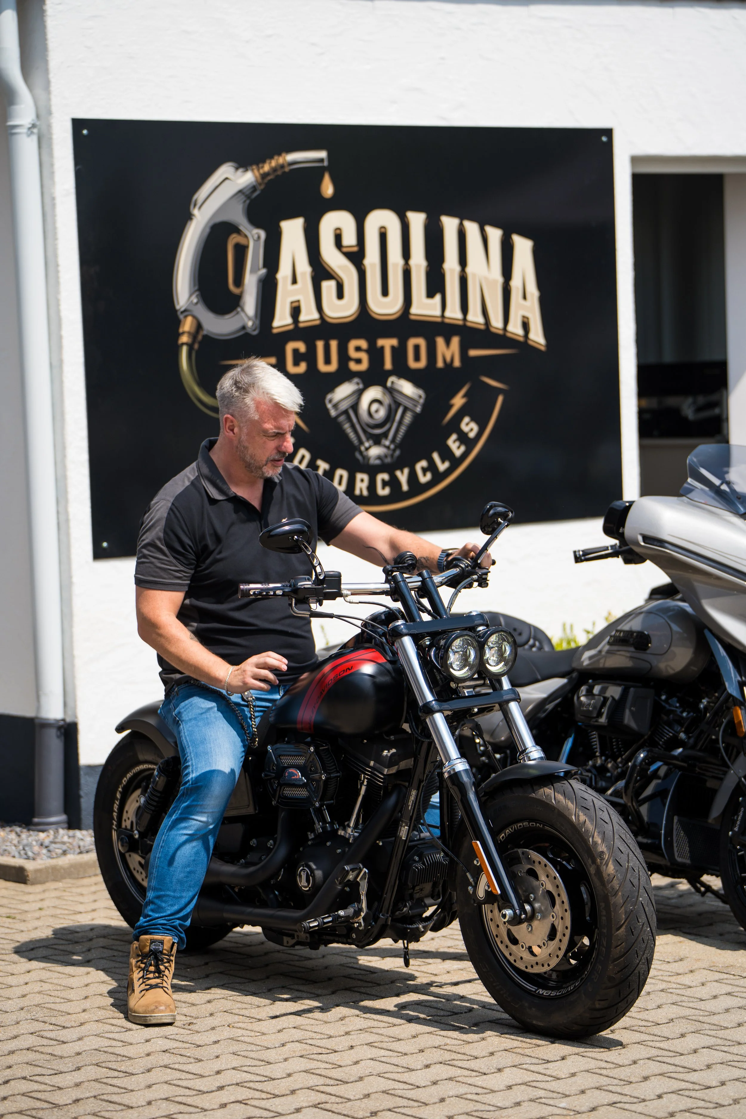 Ein Mann mit grauem Haar trägt eine schwarze T-Shirt, blaue Jeans und braune Stiefel und steht neben einer schwarzen Harley-Davidson-Motorrad vor einem Geschäft, das eine große schwarze Wand mit dem Logo "Gasolina Custom Motorcycles" hat.