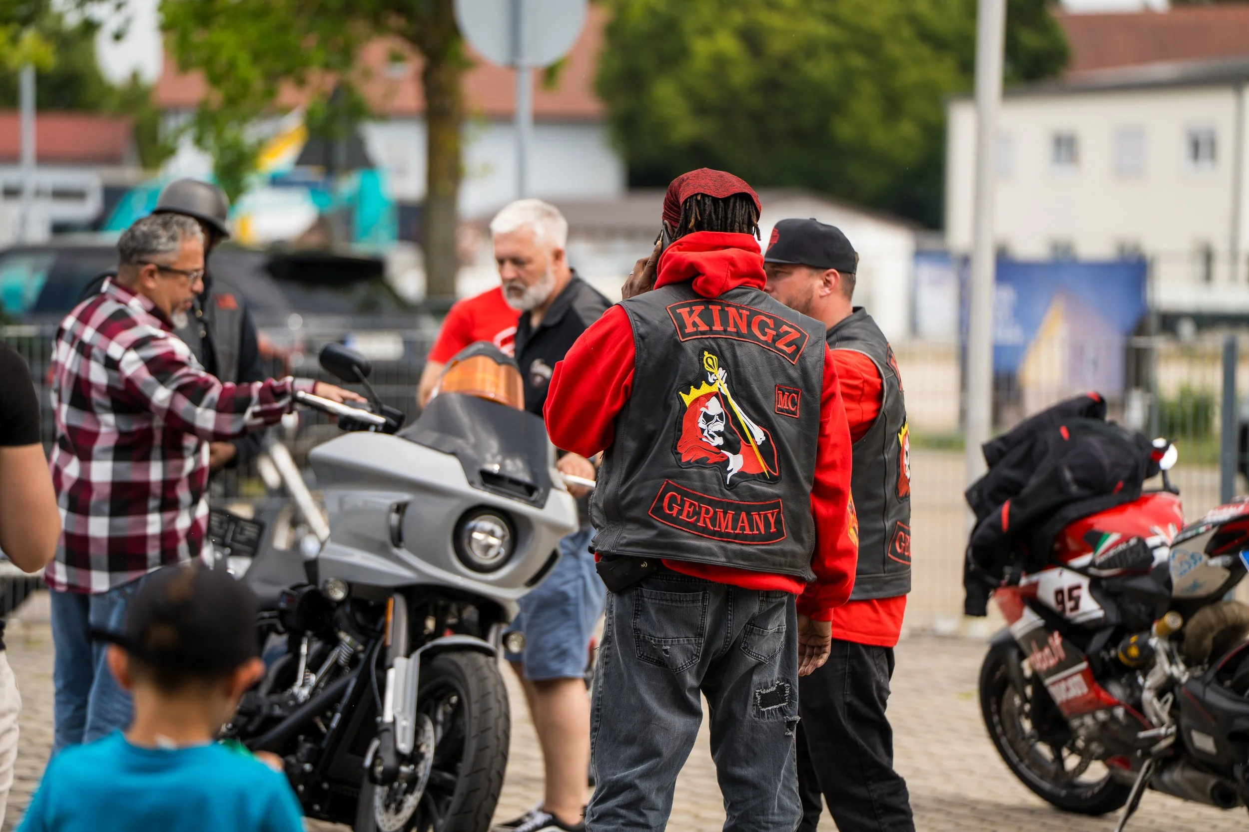 Menschen treffen sich bei einem Motorradevent im Freien. Mehrere Personen sind neben Motorrädern, einige tragen Jacken mit Aufdruck 'KINGZ GERMANY'.  Bei der Eröffnungsfeier von Gasolina Custom Motorcycles.