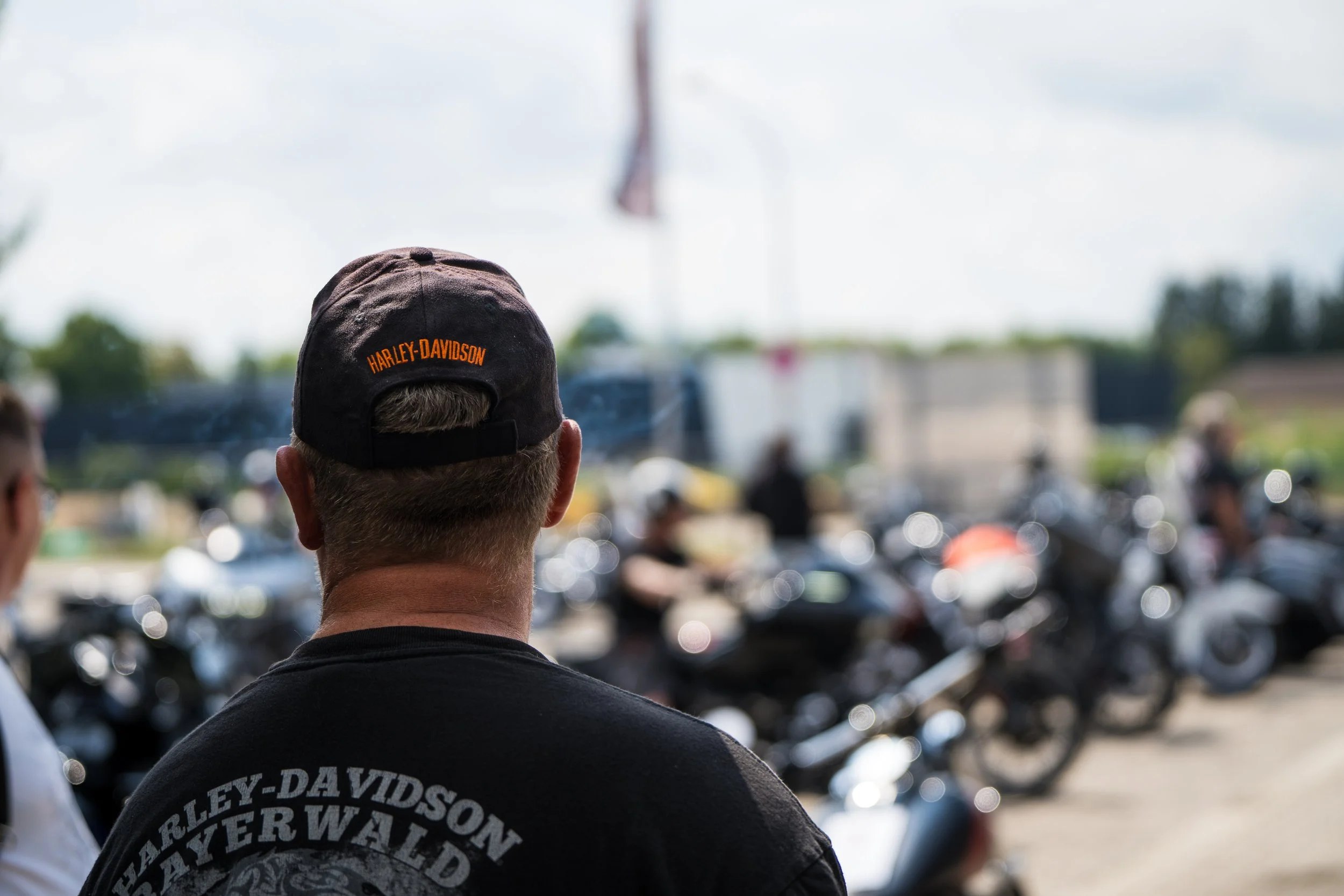 Ein Mann trägt einen schwarzen Harley-Davidson Cap und ein schwarzes T-Shirt, im Hintergrund sind Motorräder und Menschen bei der Eröffnungsfeier von Gasolina Custom Motorcycles, die im Freien bei sonnigem Himmel stattfindet.  