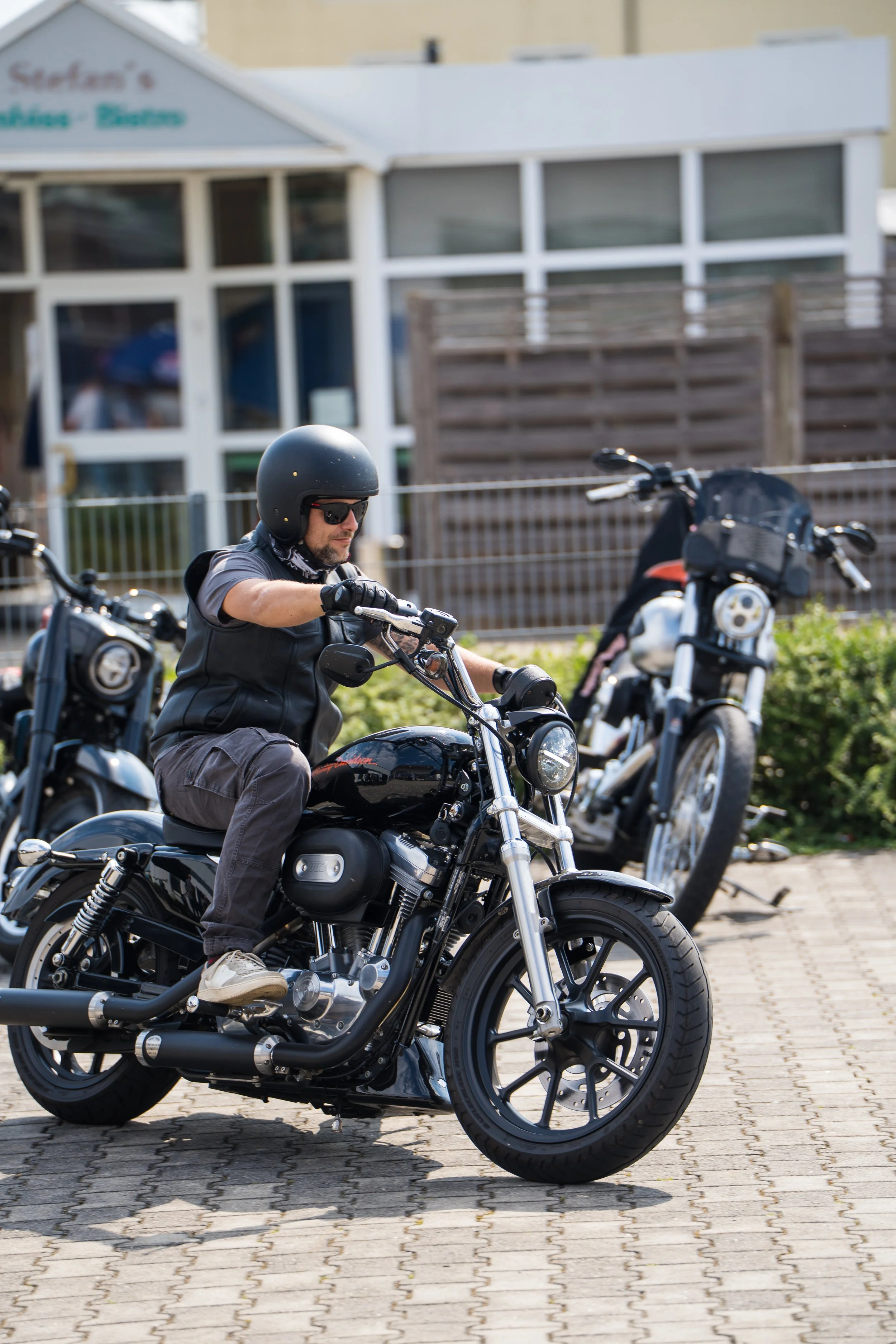 Ein Mann trägt Helm und Sonnenbrille, fährt Motorrad, steht auf gepflastertem Platz von Gasolina Custom Motorcycles, im Hintergrund Mehrfamilienhaus und Holzsperrzaun.