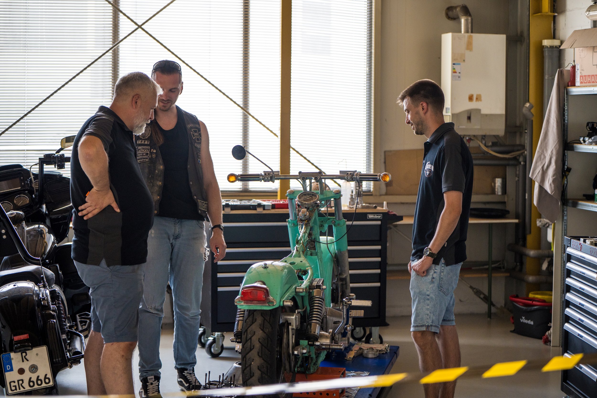 Vier Männer in einer Werkstatt arbeiten an einem Motorcycle. Zwei von ihnen schauen auf den Motor, während die anderen beiden daneben stehen.  Bei der Eröffnungsfeier von Gasolina Custom Motorcycles.
