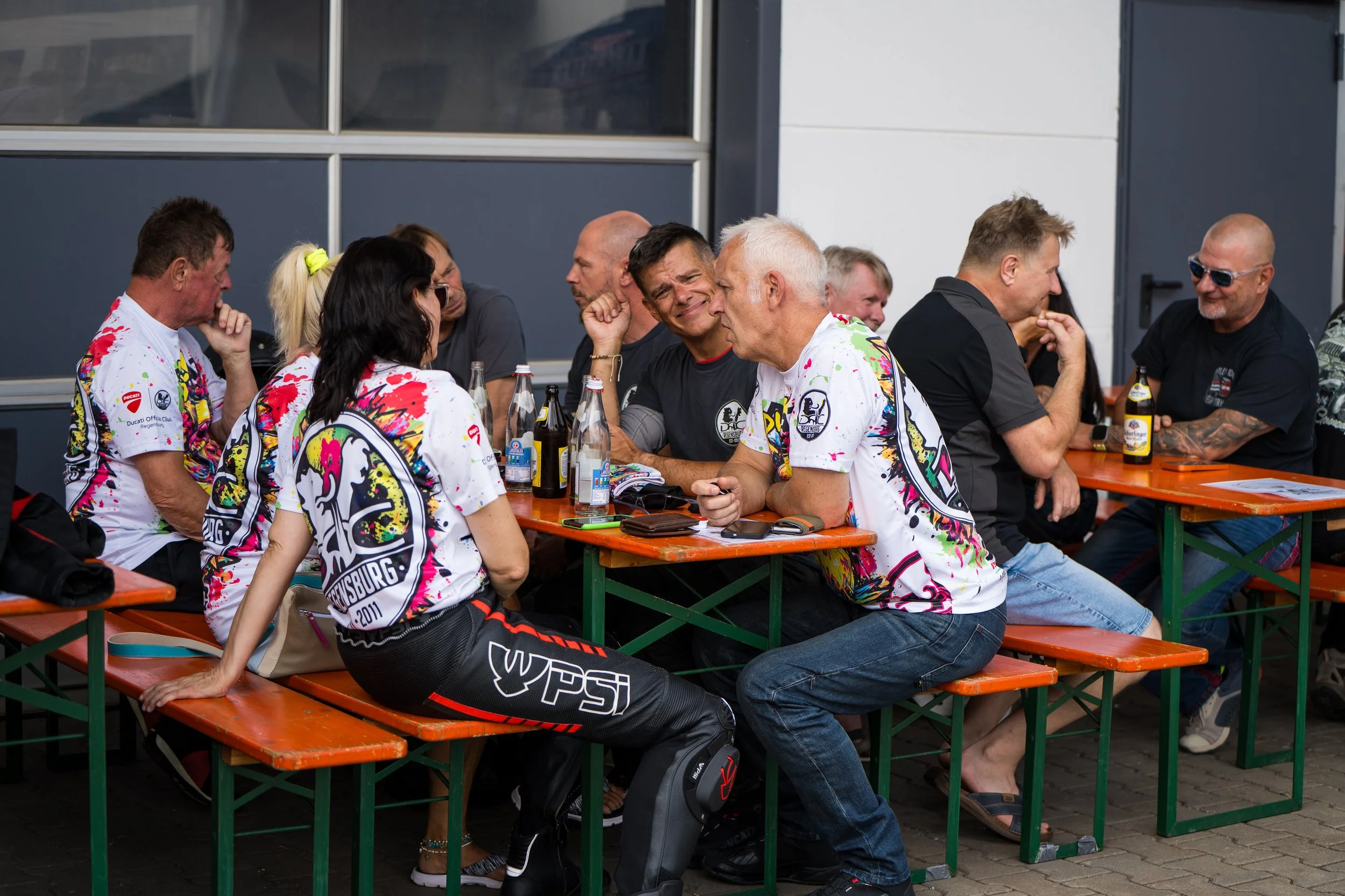 Eine Gruppe von Menschen sitzt an einem orangefarbenen Holztisch im Freien, sie scheinen sich zu unterhalten.  einige Getränke stehen auf dem Tisch. Bei der Eröffnungsfeier von Gasolina Custom Motorcycles.
