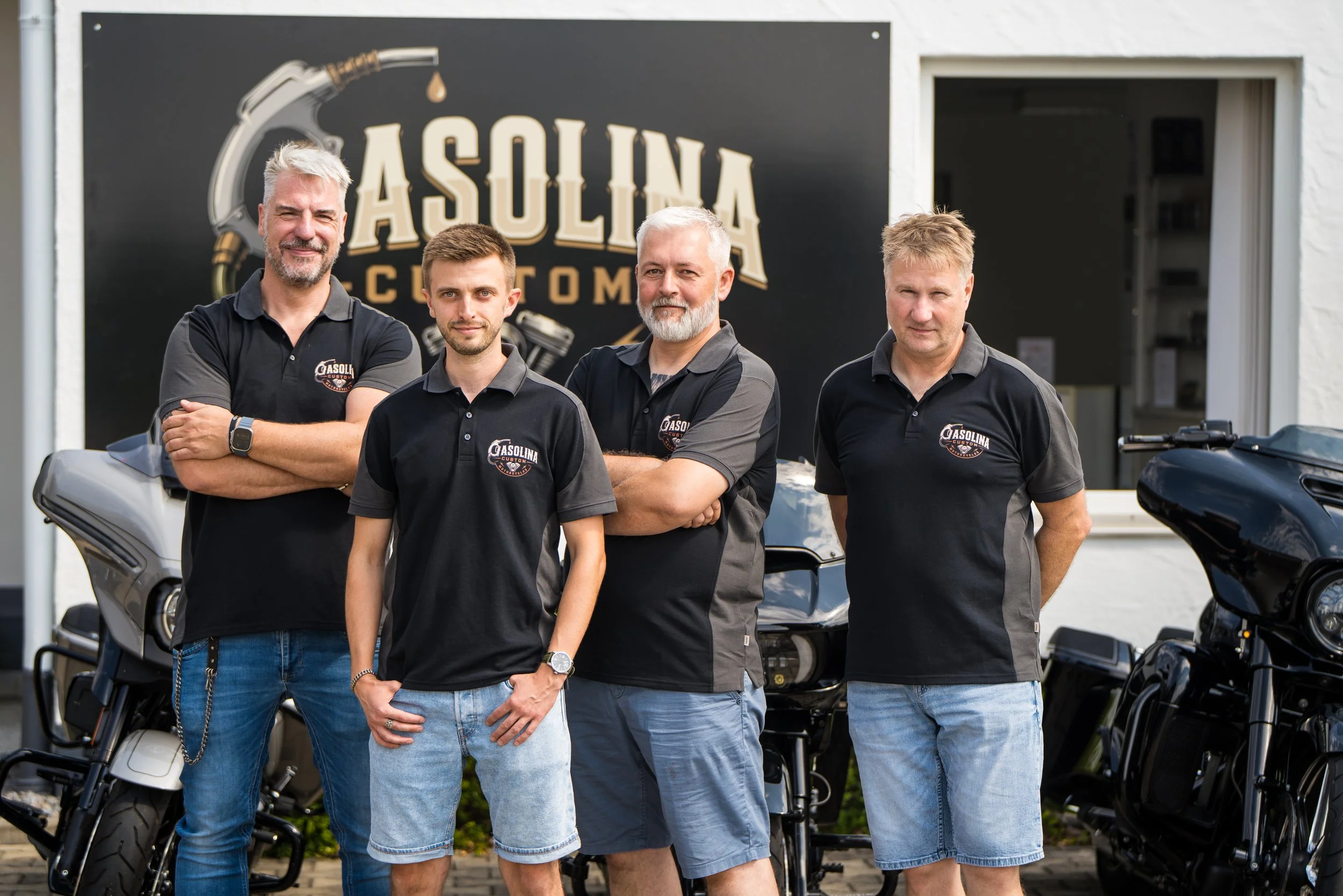 Das Team von Gasolina Custom Motorcycles. Sie tragen schwarze T-Shirts mit demselben Logo, stehen nebeneinander, mit Motorrädern im Vordergrund.