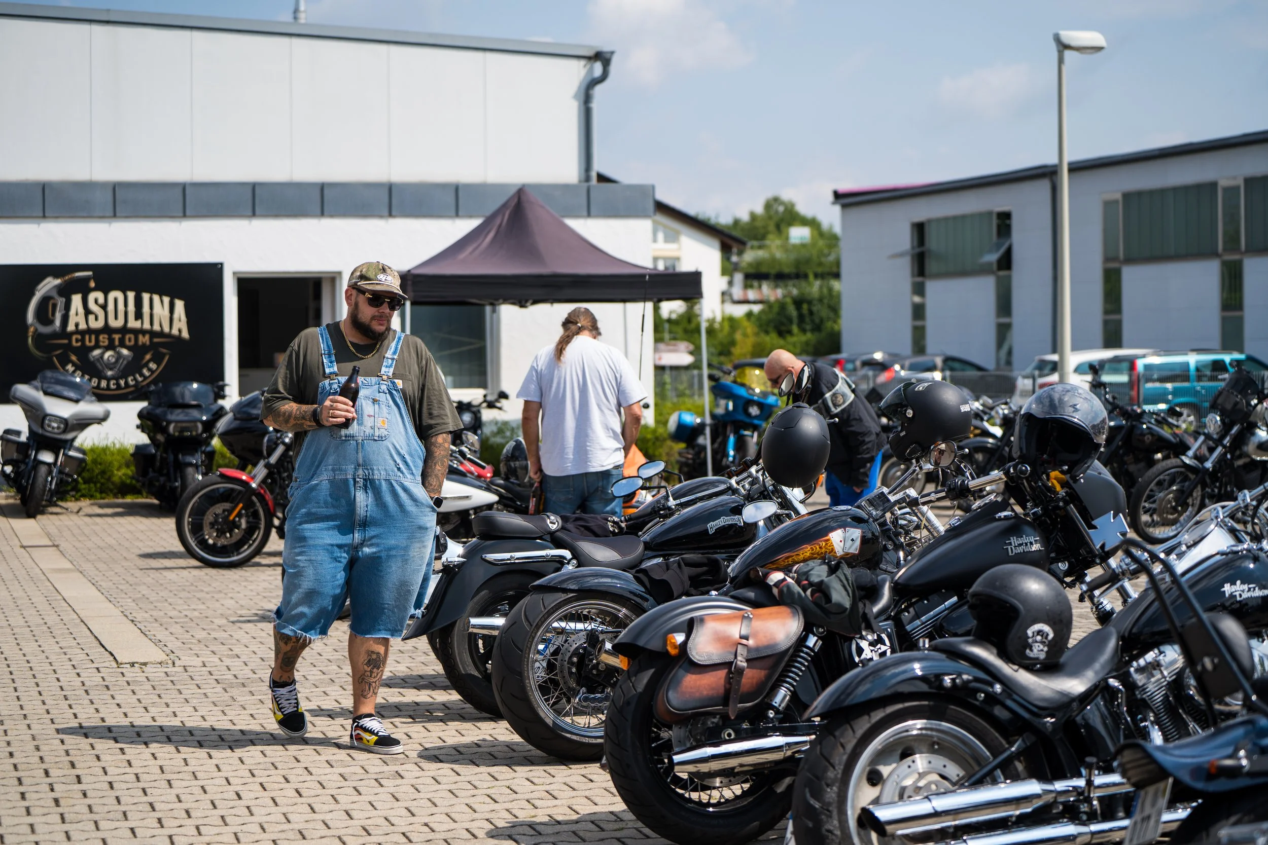 Menschen stehen in einer Motorradparkhalle, einige betrachten die Motorräder, im Hintergrund befinden sich moderne Gebäude und ein blauer Himmel.  Bei der Eröffnungsfeier von Gasolina Custom Motorcycles.