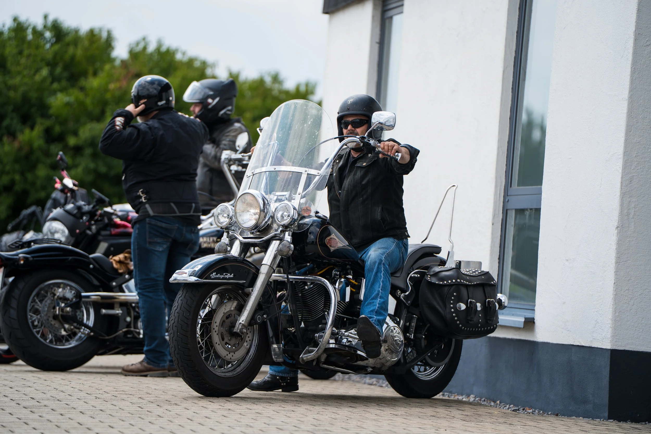 Mehrere Motorradfahrer, die Helme tragen, vor einem modernen Gebäude, einer davon sitzt auf einem schwarzen Harley-Davidson Motorrad mit Windschutzscheibe. Bei der Eröffnungsfeier von Gasolina Custom Motorcycles.