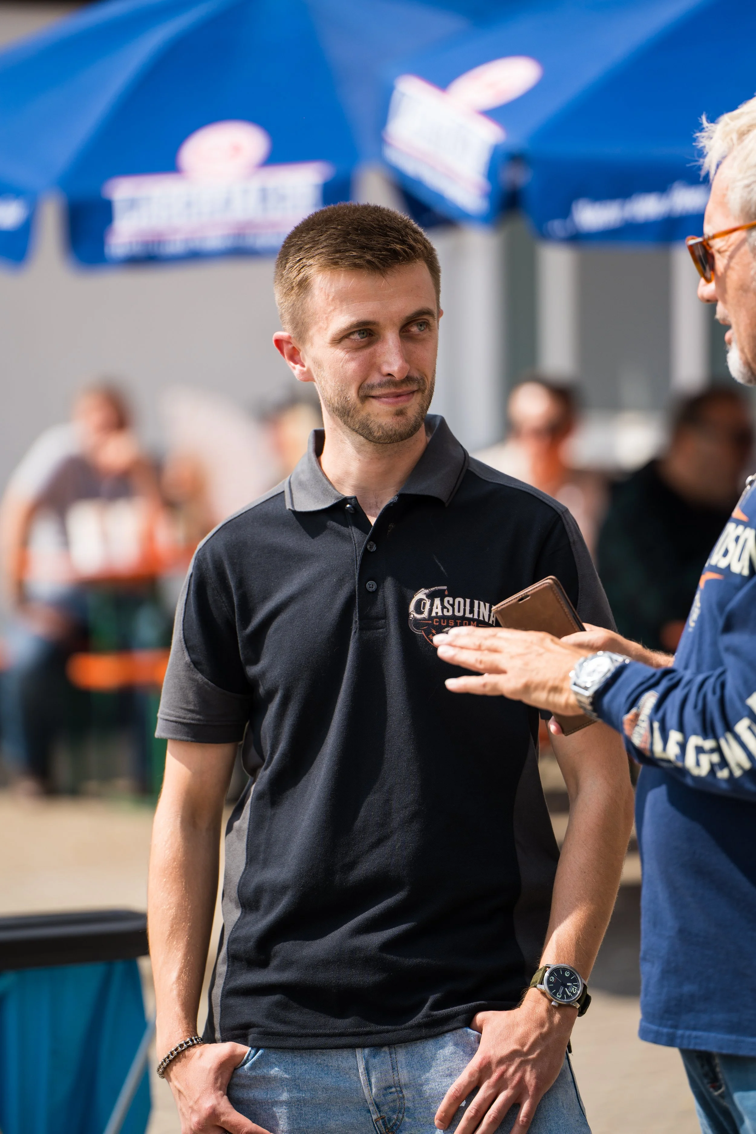 Junger Mann im schwarzen T-Shirt spricht mit älterem Mann unter einem blauen Sonnenschirm, im Hintergrund sind weitere Personen sichtbar. Bei der Eröffnungsfeier von Gasolina Custom Motorcycles.