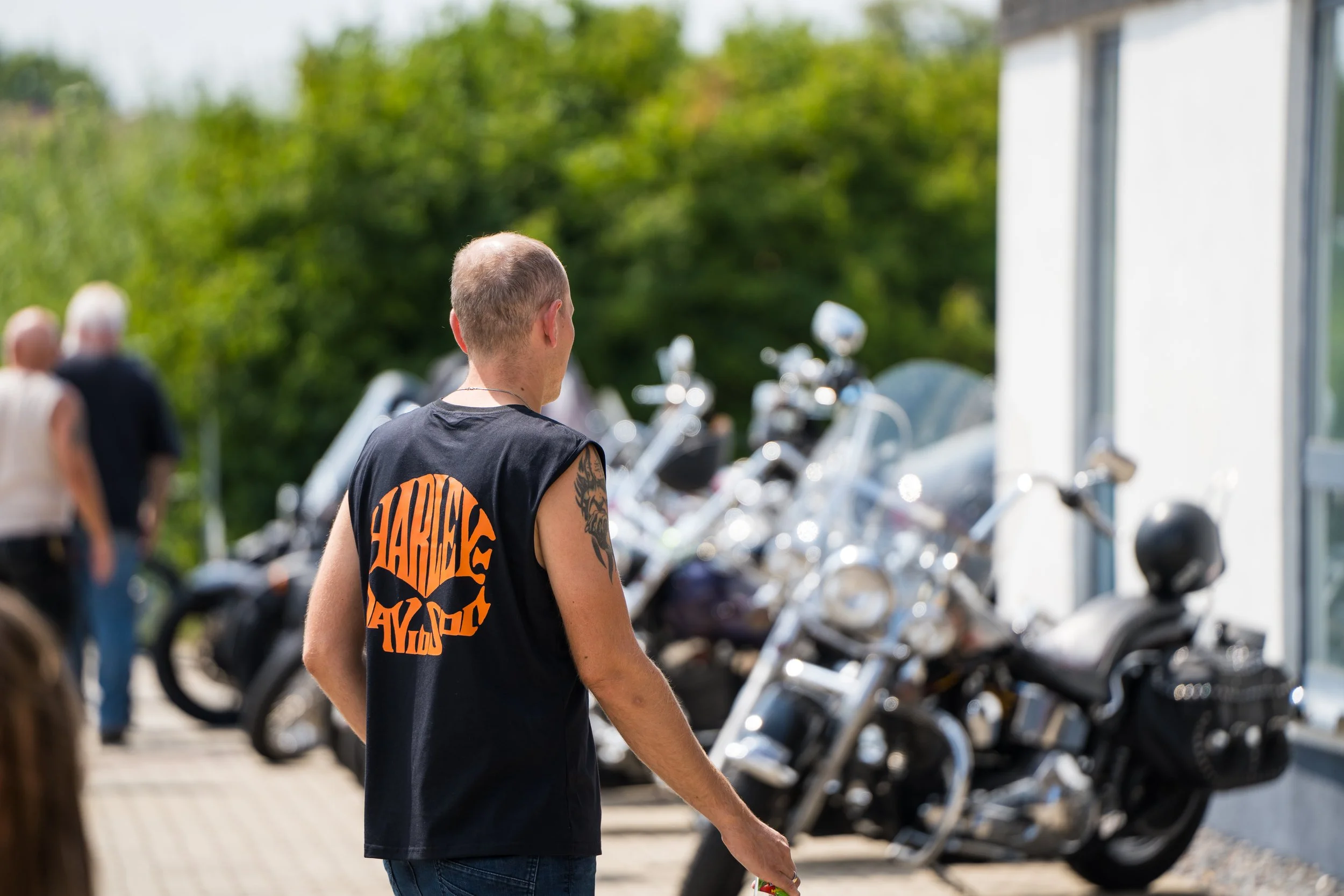 Junger Mann mit kurzen Haaren und Tattoos am Arm steht vor einer Reihe von abgestellten Motorrädern, im Hintergrund sind andere Menschen und grüne Bäume.  Bei der Eröffnungsfeier von Gasolina Custom Motorcycles.